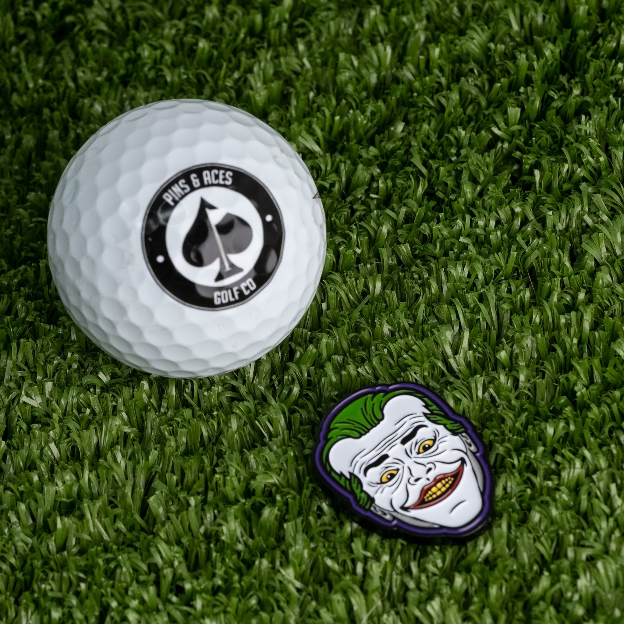 Joker Ball Marker 3.0 next to a golf ball featuring the Pins and Aces logo on a green turf background.