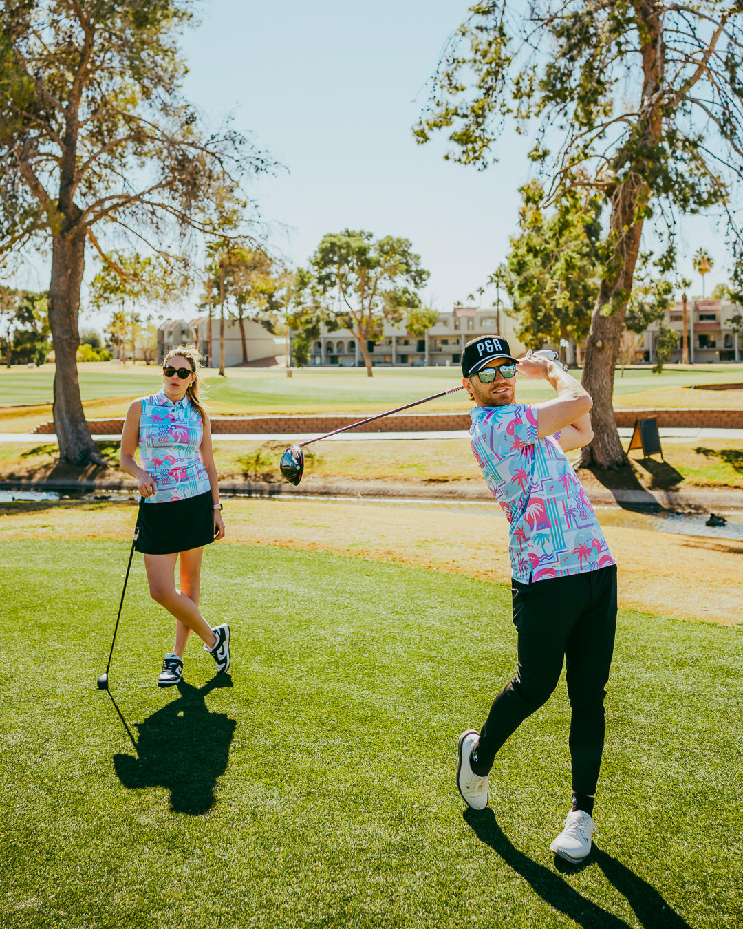 A man and woman in vibrant beach-themed golf apparel swing clubs on a sunny golf course, surrounded by palm trees.