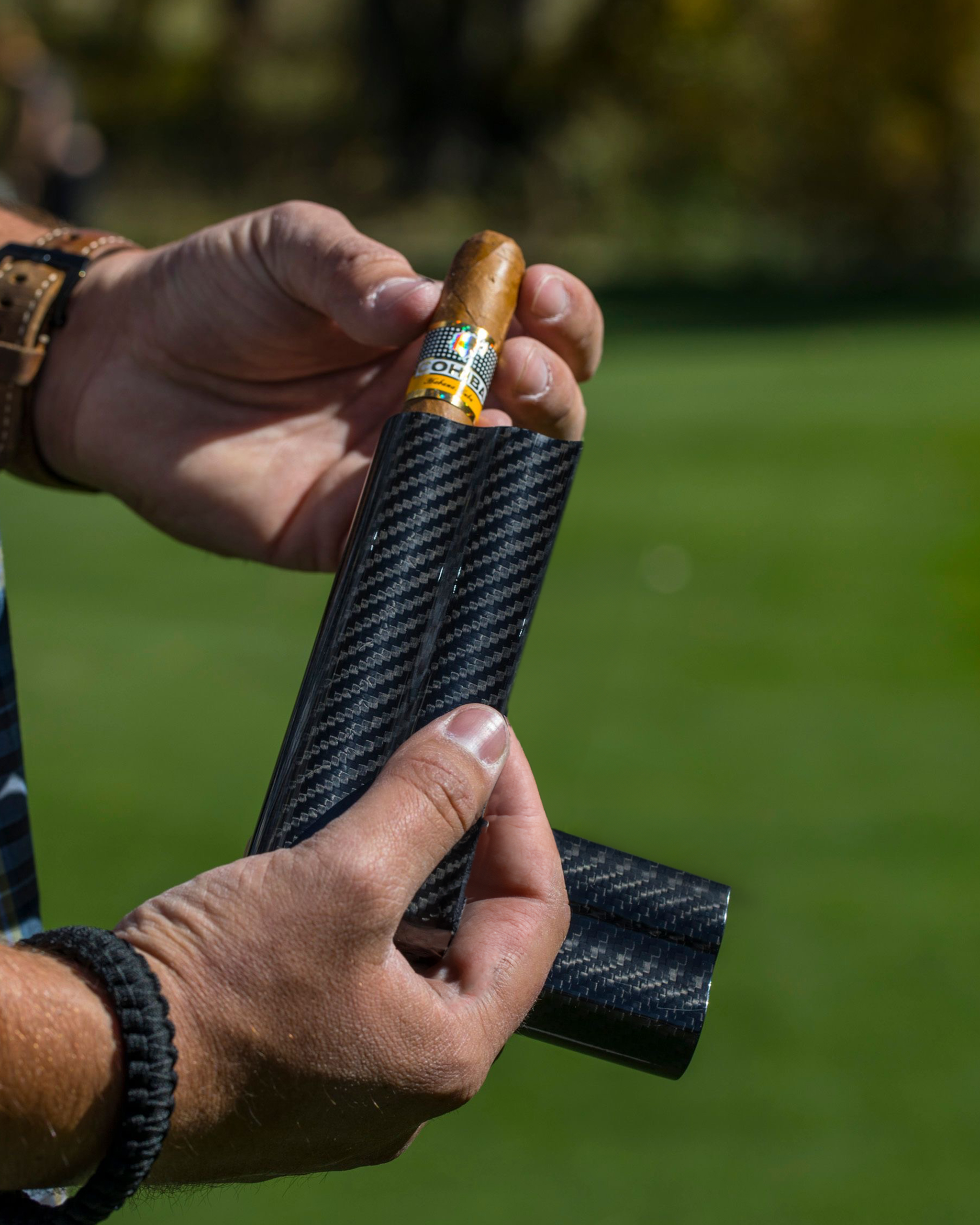 Close-up of a hand placing a cigar into a black carbon fiber golf club headcover, outdoors on a sunny day.