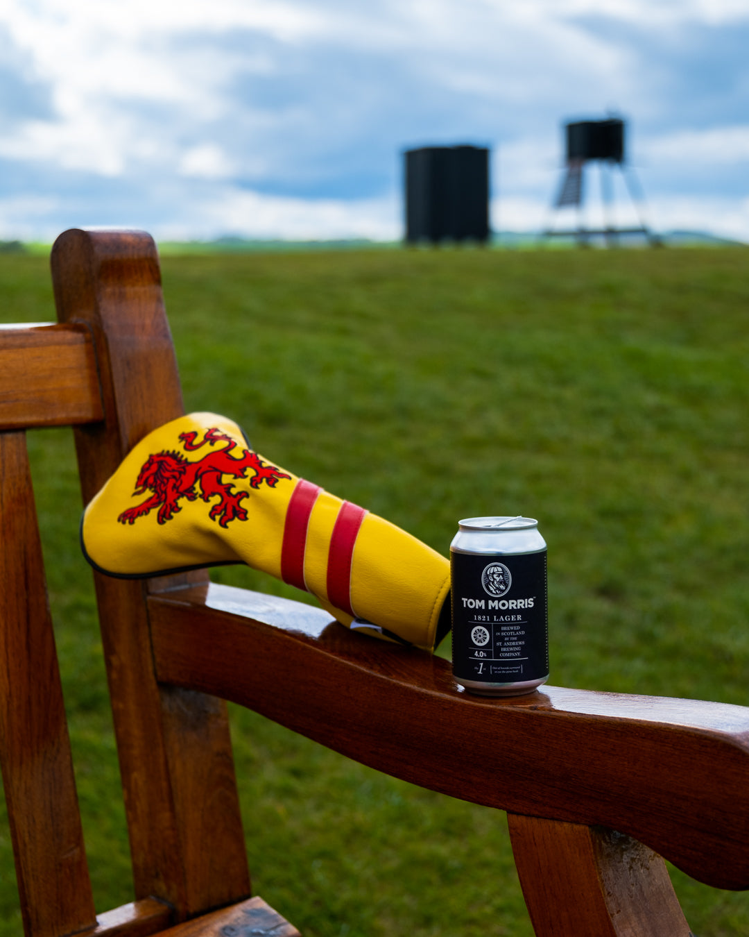Scotland driver cover with a lion design resting on a wooden chair beside a can of Tom Morris beer on a grassy field.