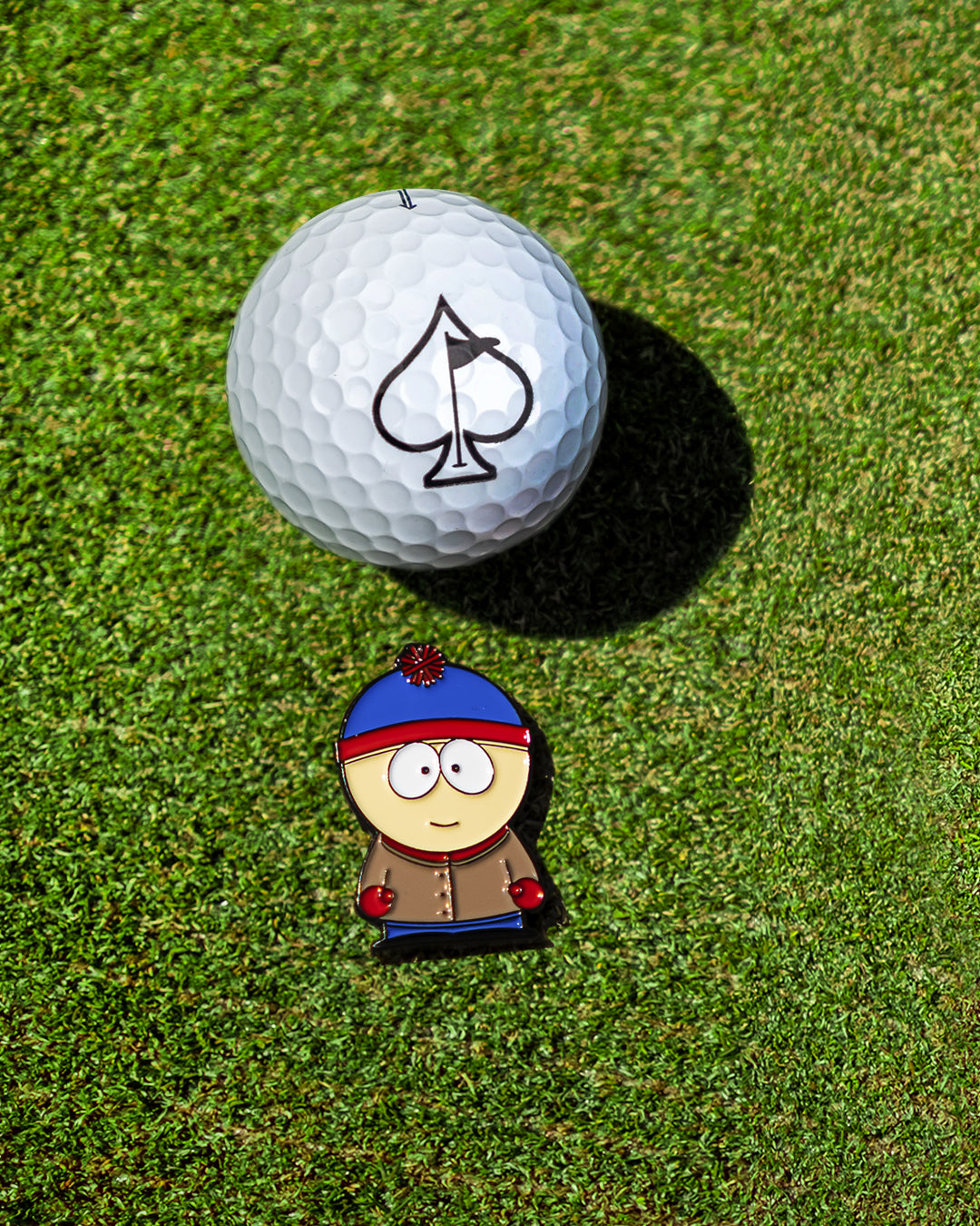 South Park Stan ball marker beside a golf ball with an ace of spades logo on a green golf course.