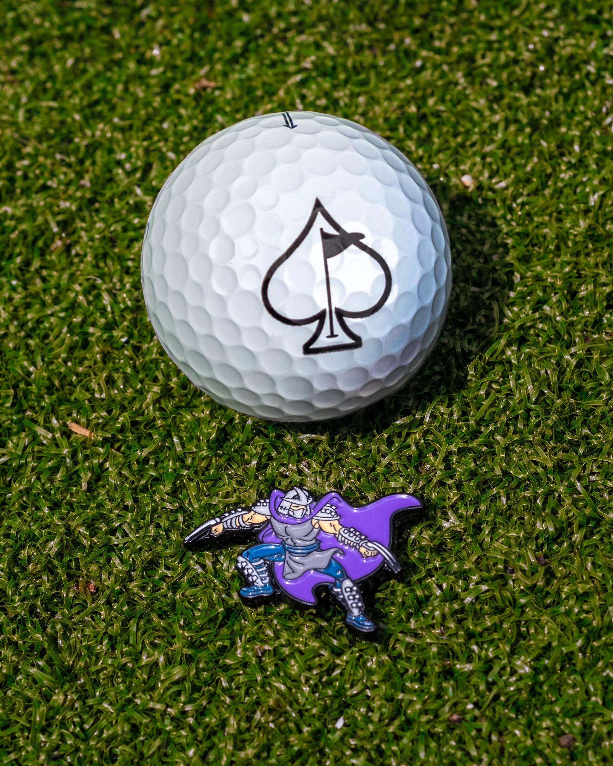 TMNT Shredder ball marker next to a golf ball with an ace of spades logo on a green turf background.