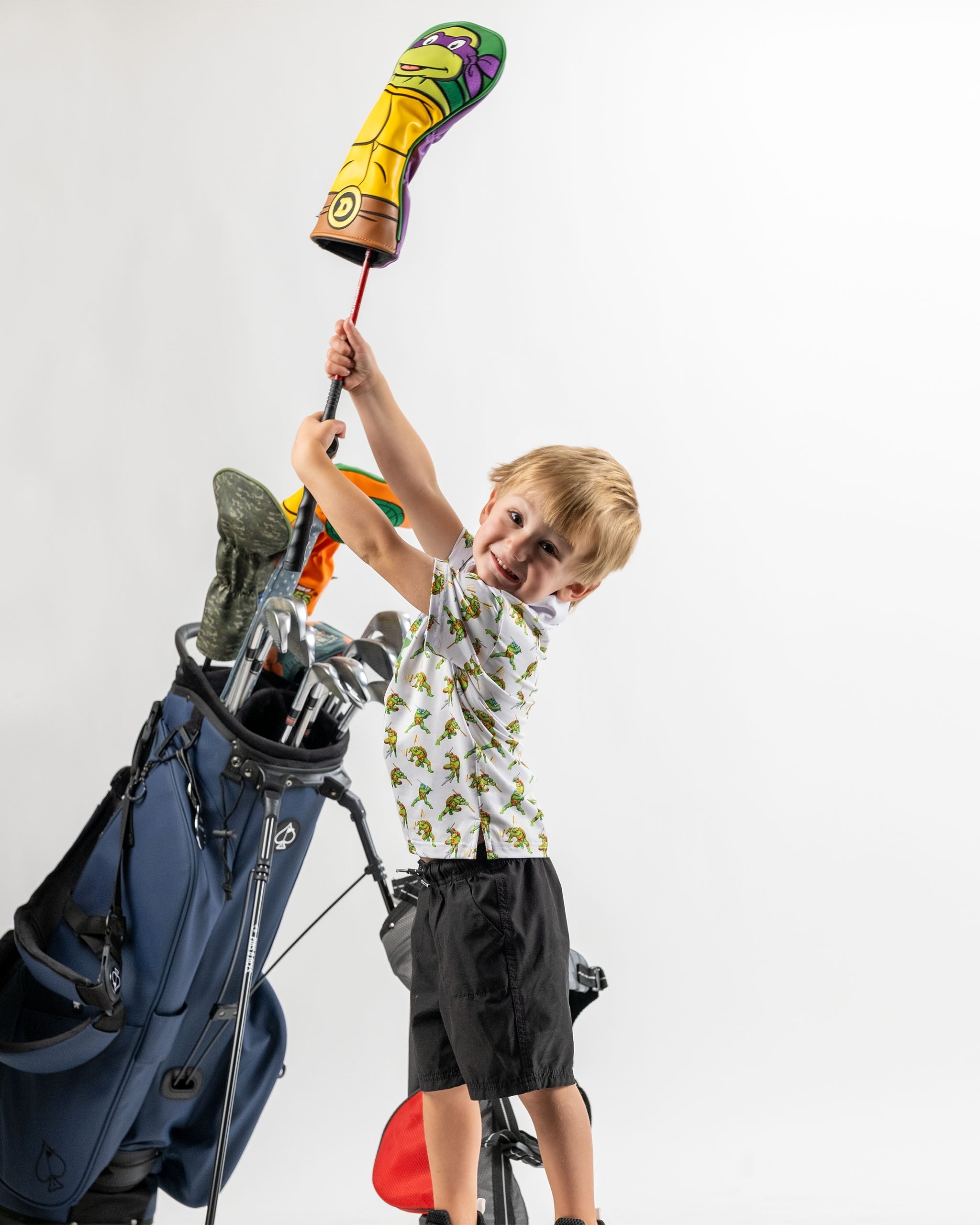 Youth holding a TMNT Cowabunga Classic 8-Bit headcover while posing next to a golf bag, showcasing fun golf accessories.