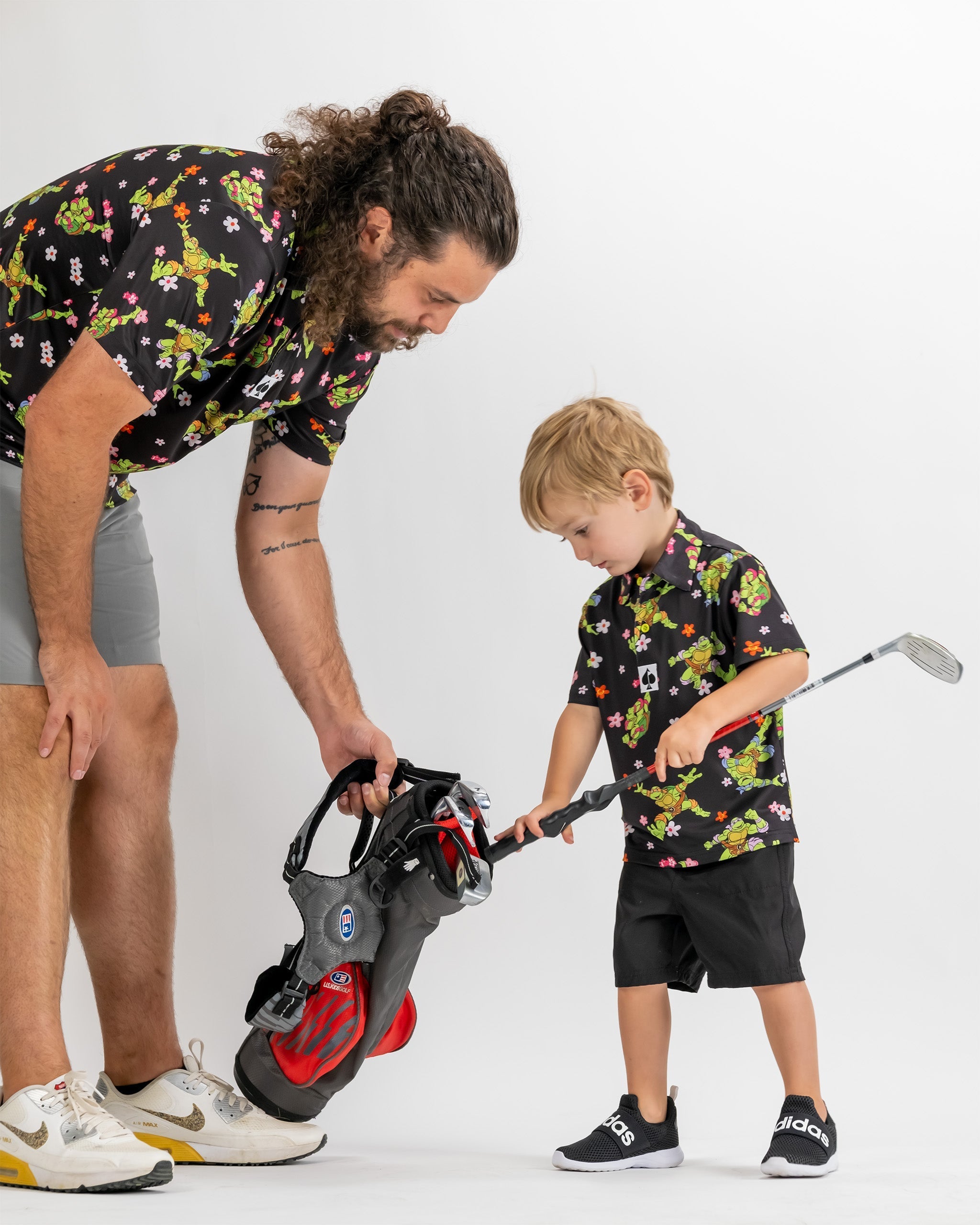A father and son in matching TMNT floral shirts, with the son holding a golf club and inspecting a golf bag.