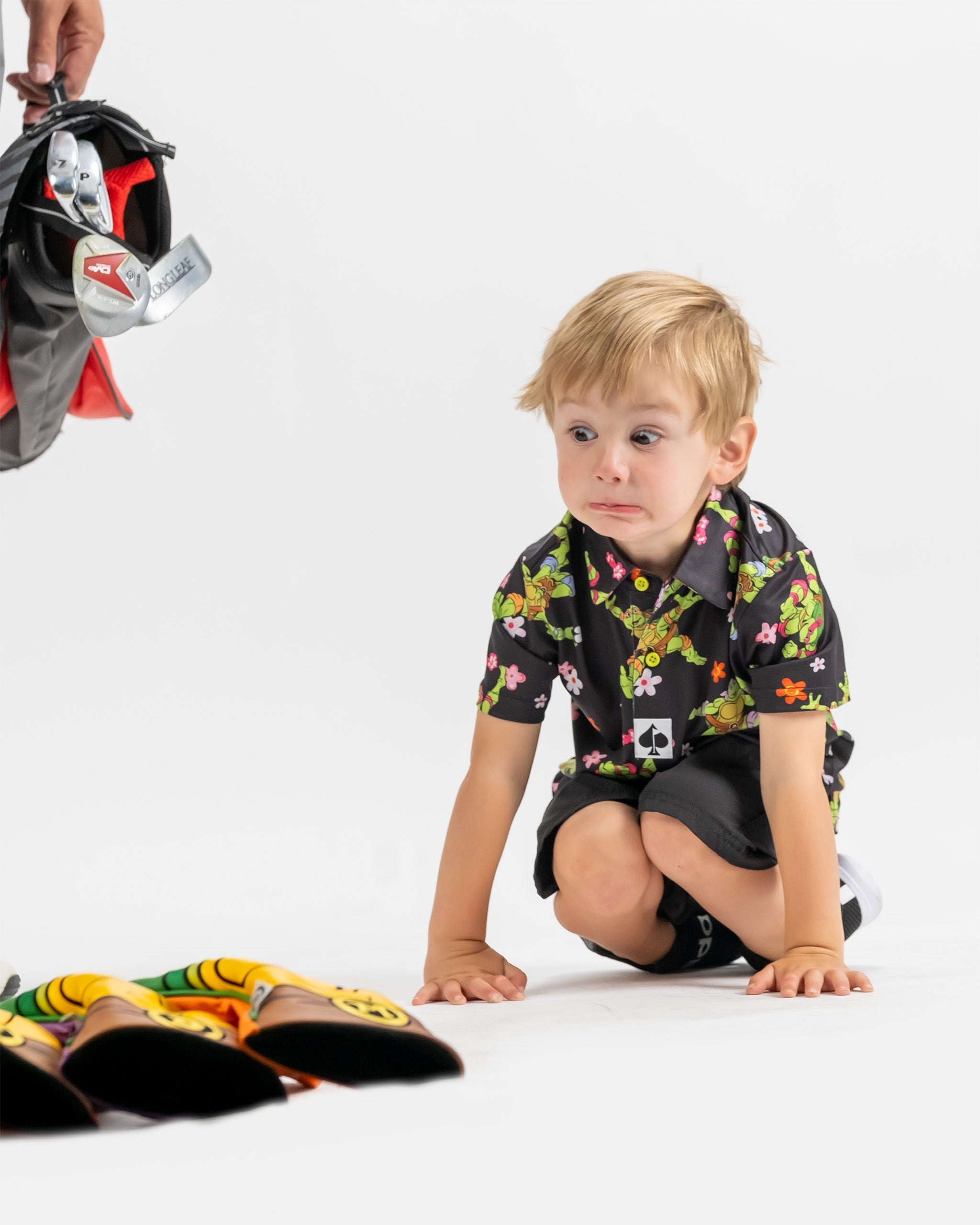 Youth wearing a black floral TMNT shirt, kneeling on the floor beside colorful golf headcovers, looking surprised.