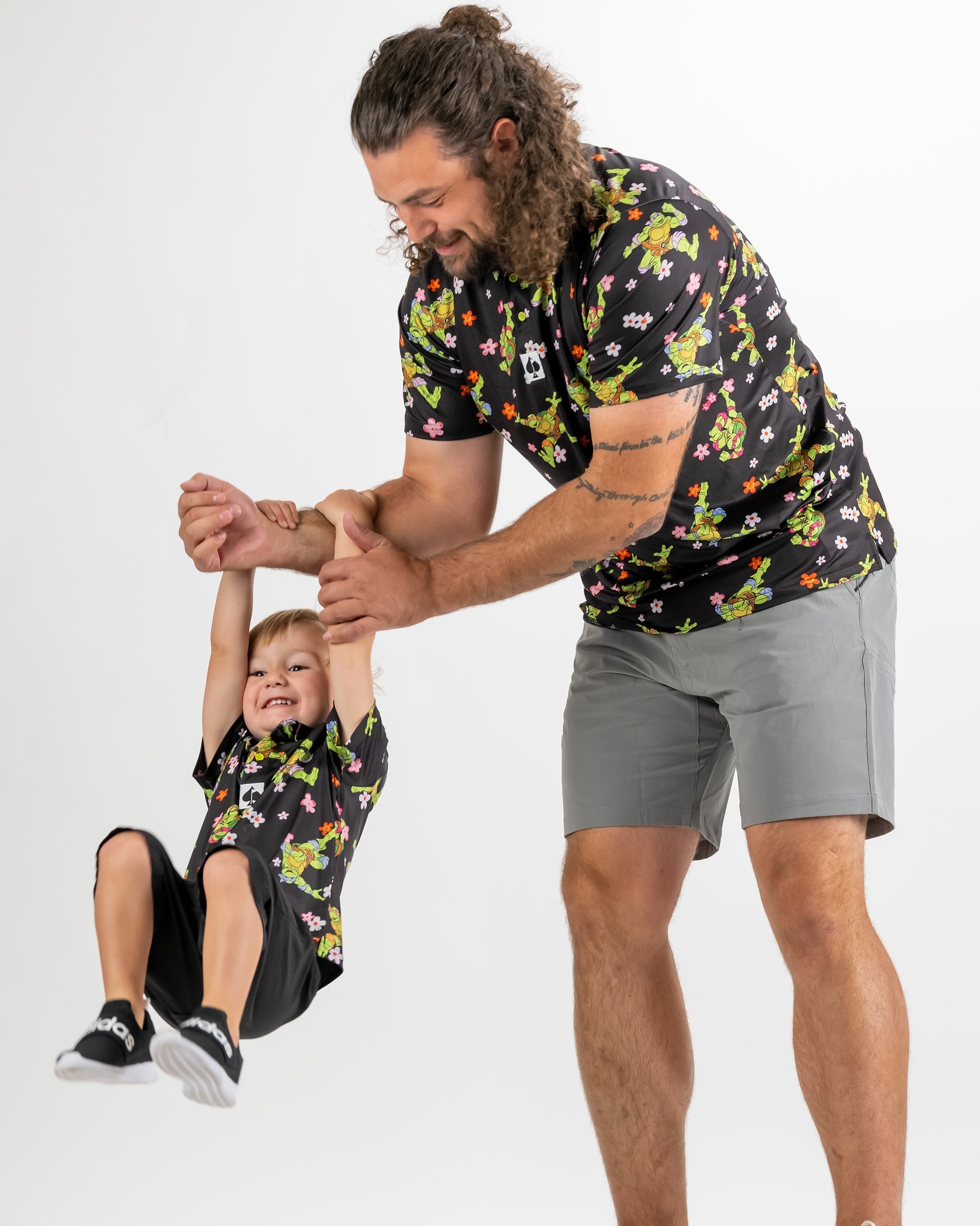 A father lifts his smiling son while both wear matching TMNT flower-patterned shirts, showcasing a fun family moment.