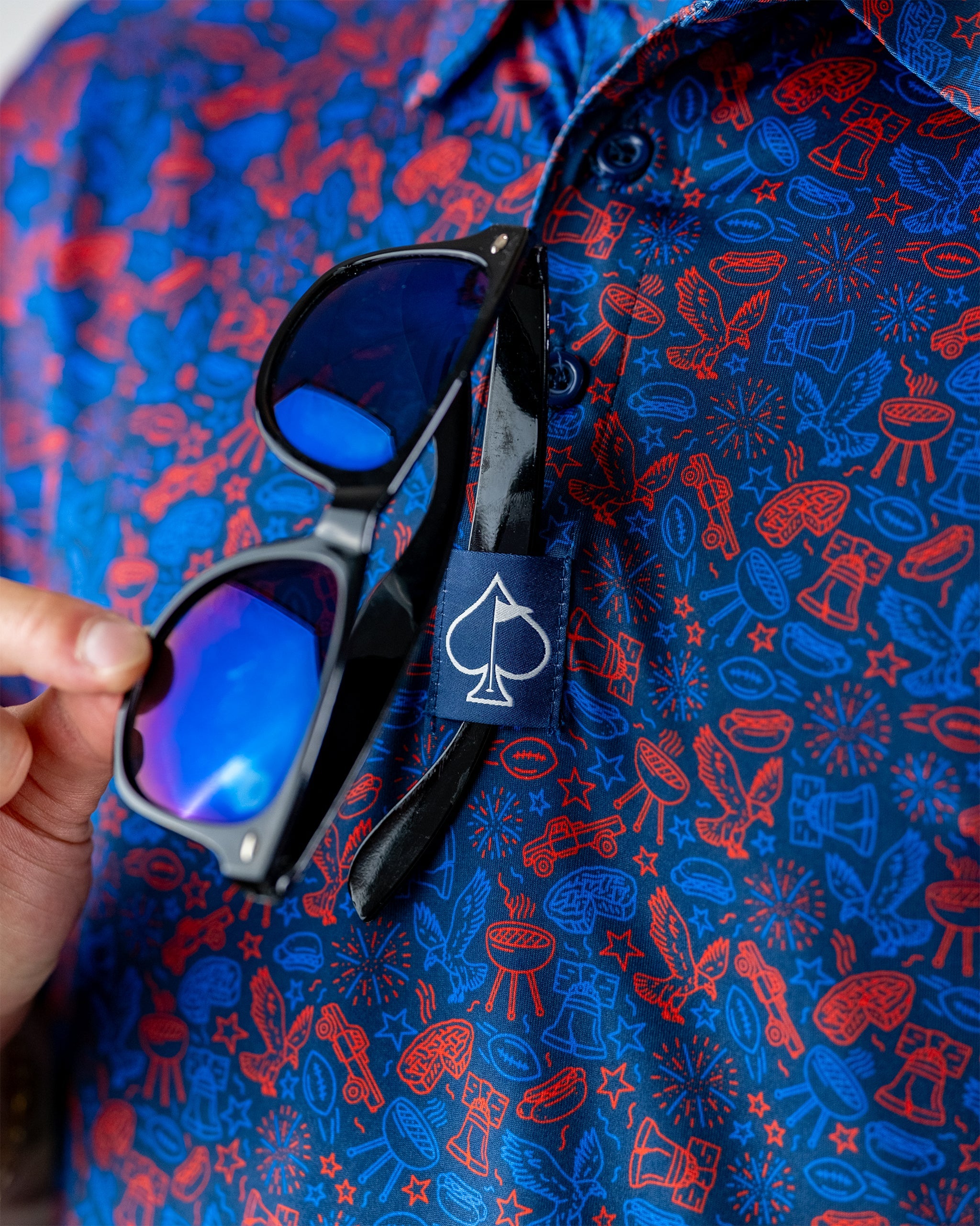 Navy shirt with a colorful doodle pattern featuring fireworks and golf motifs, holding sunglasses with a logo tag.