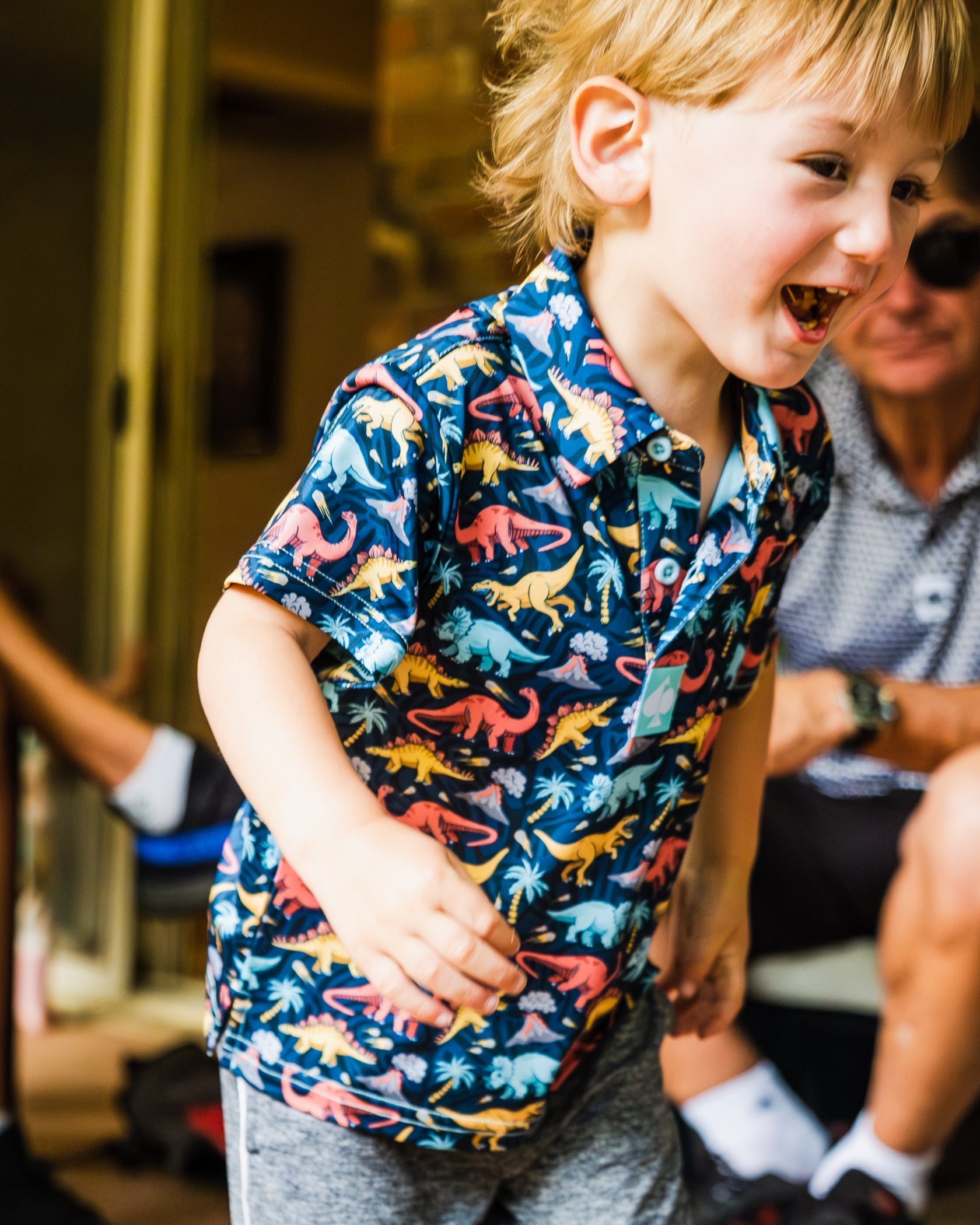 A young boy joyfully running in a colorful Jurassic-themed polo shirt featuring dinosaurs and tropical designs.