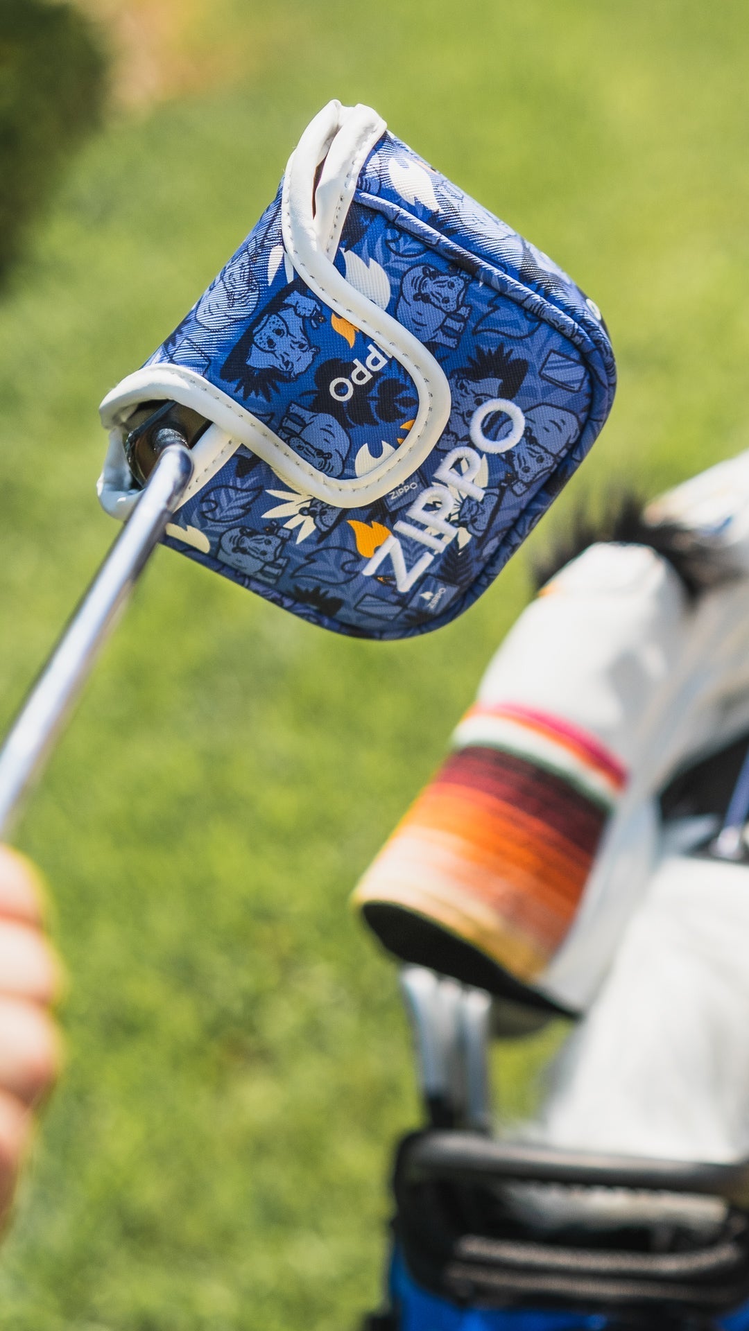 Bold blue Zippo mallet putter cover featuring a graphic design, held above a golf bag on a green course.