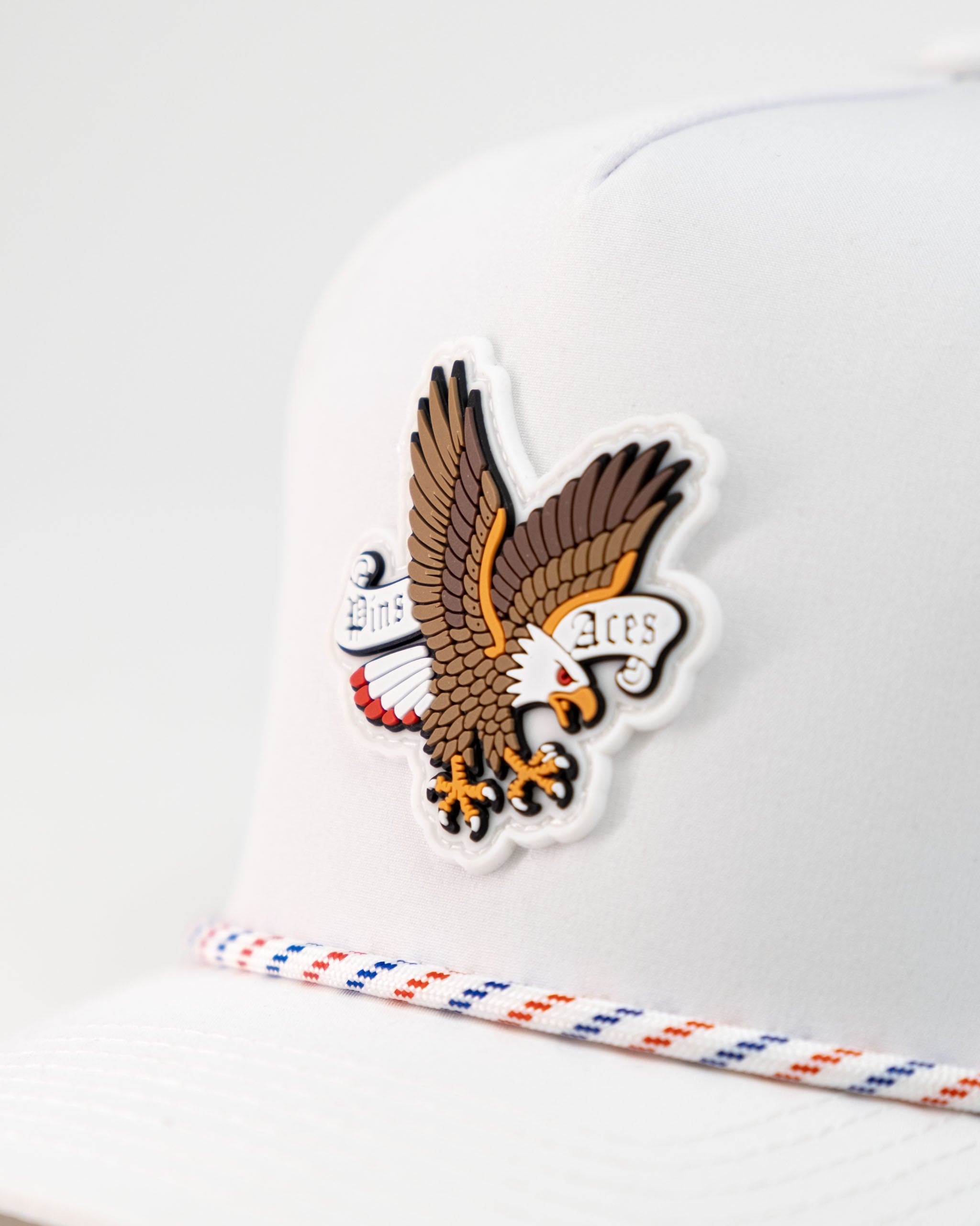 White perforated rope hat featuring a vibrant eagle graphic and "Pins and Aces" branding for stylish golf enthusiasts.
