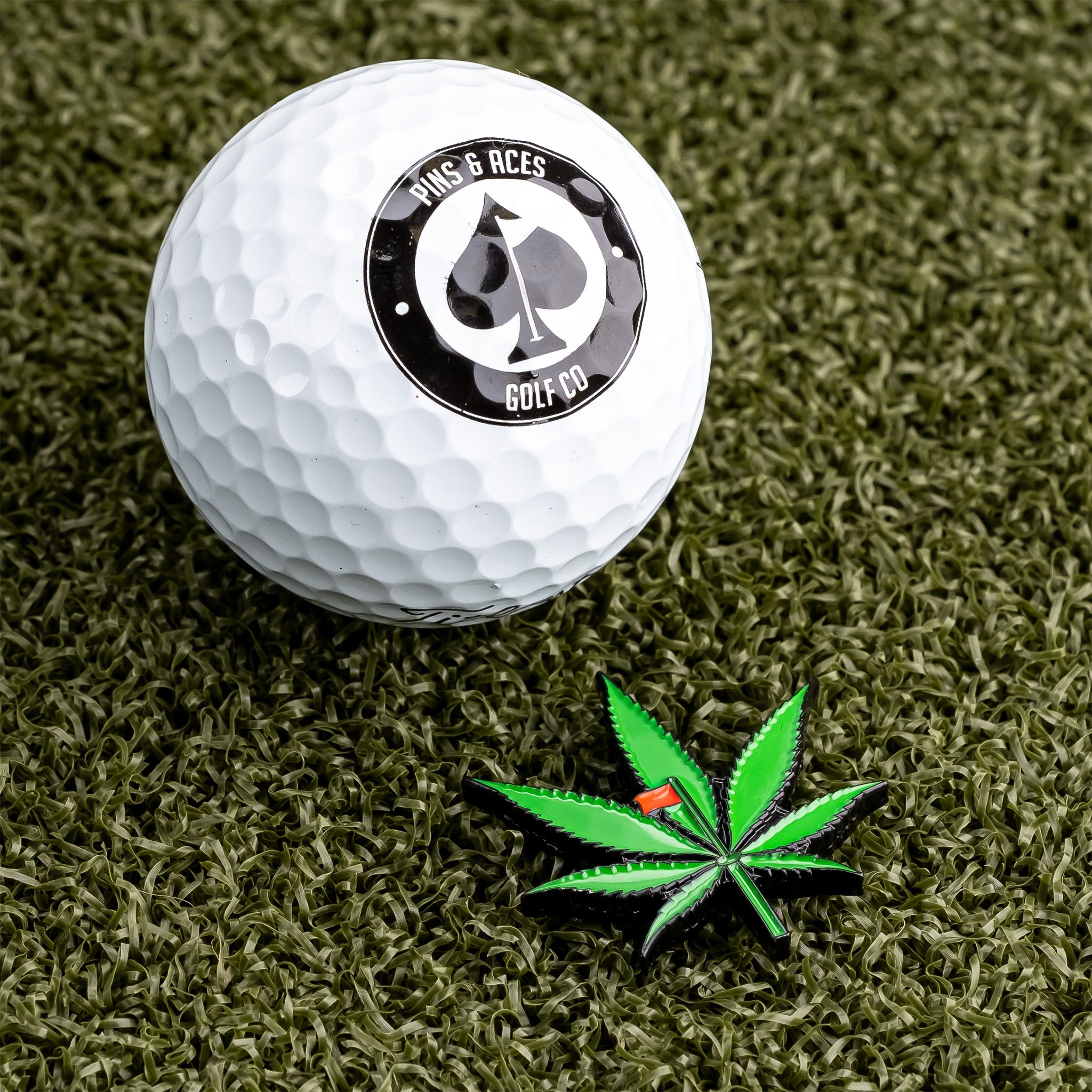 Green pot leaf ball marker on a golf ball, surrounded by grass.