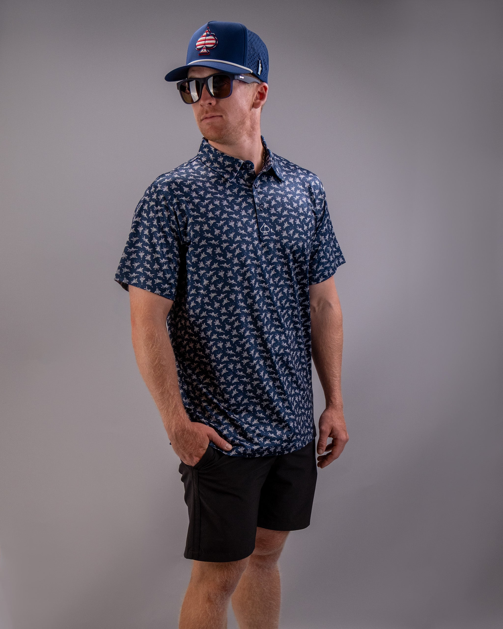 A man wearing a blue patterned short-sleeve shirt, black shorts, a blue baseball cap with a logo, sunglasses, and standing against a plain gray background.