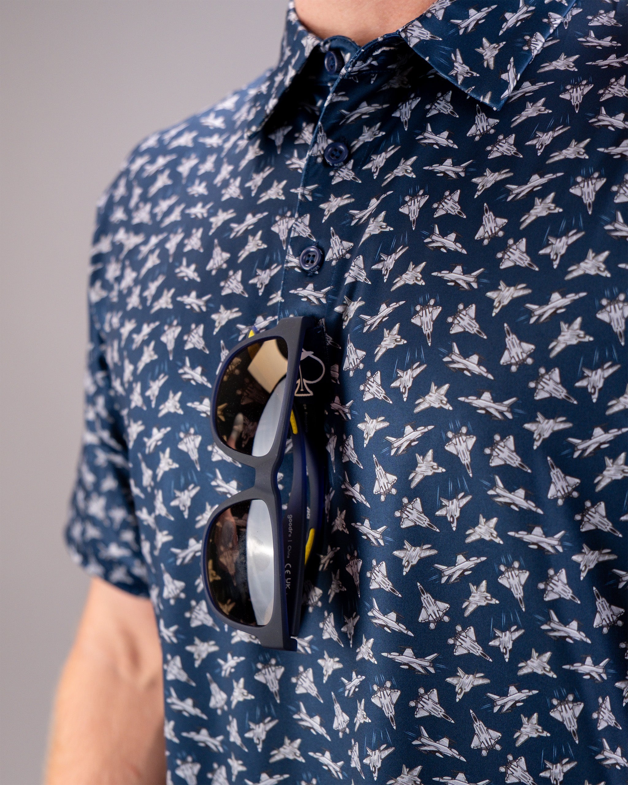 Close-up of a person wearing a navy blue shirt with a pattern of fighter jets, with sunglasses hanging from the shirt pocket.