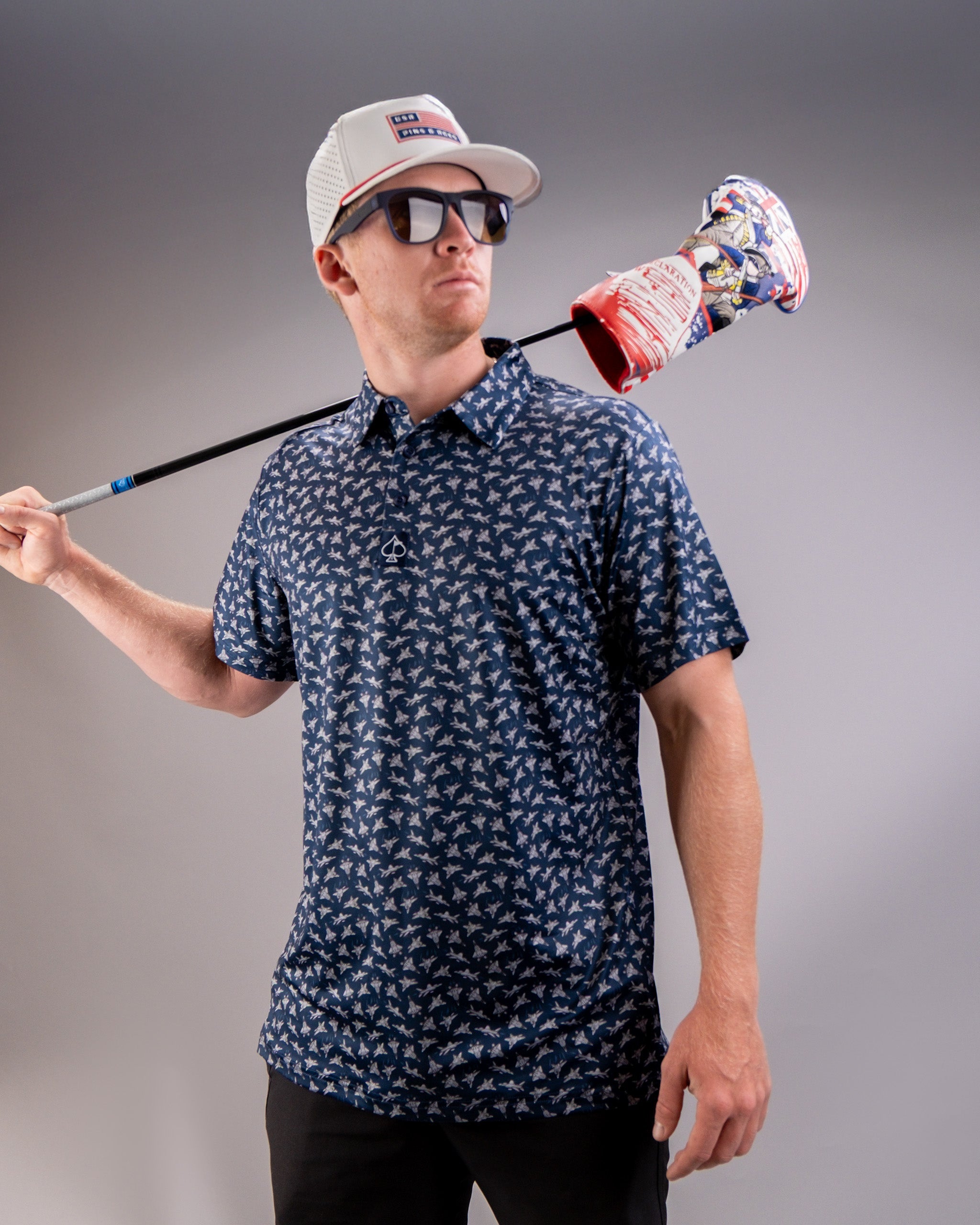 A man wearing sunglasses, a patterned shirt, and a cap carries a golf club with a colorful headcover over his shoulder.