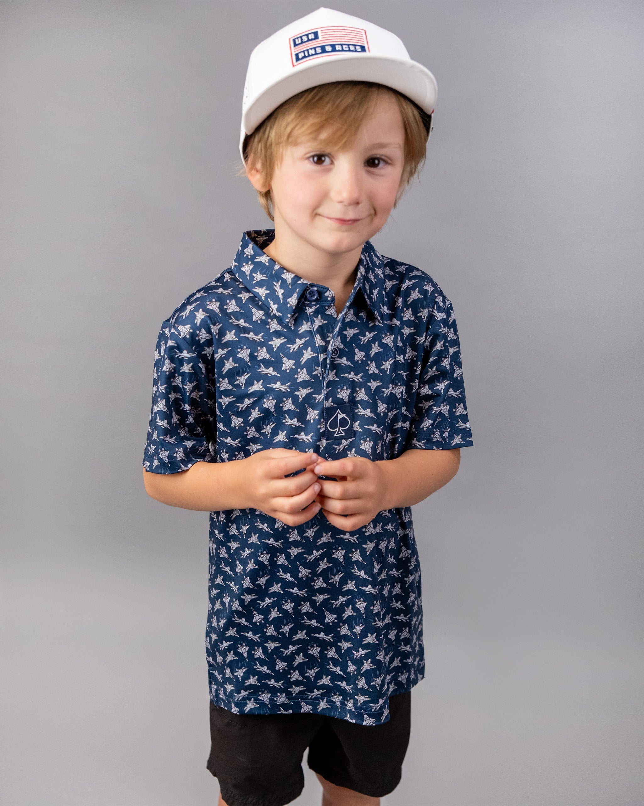 A young boy wearing a white baseball cap with a USA Pins and Aces logo, a navy blue shirt with a bird pattern, and black shorts, standing against a gray background.