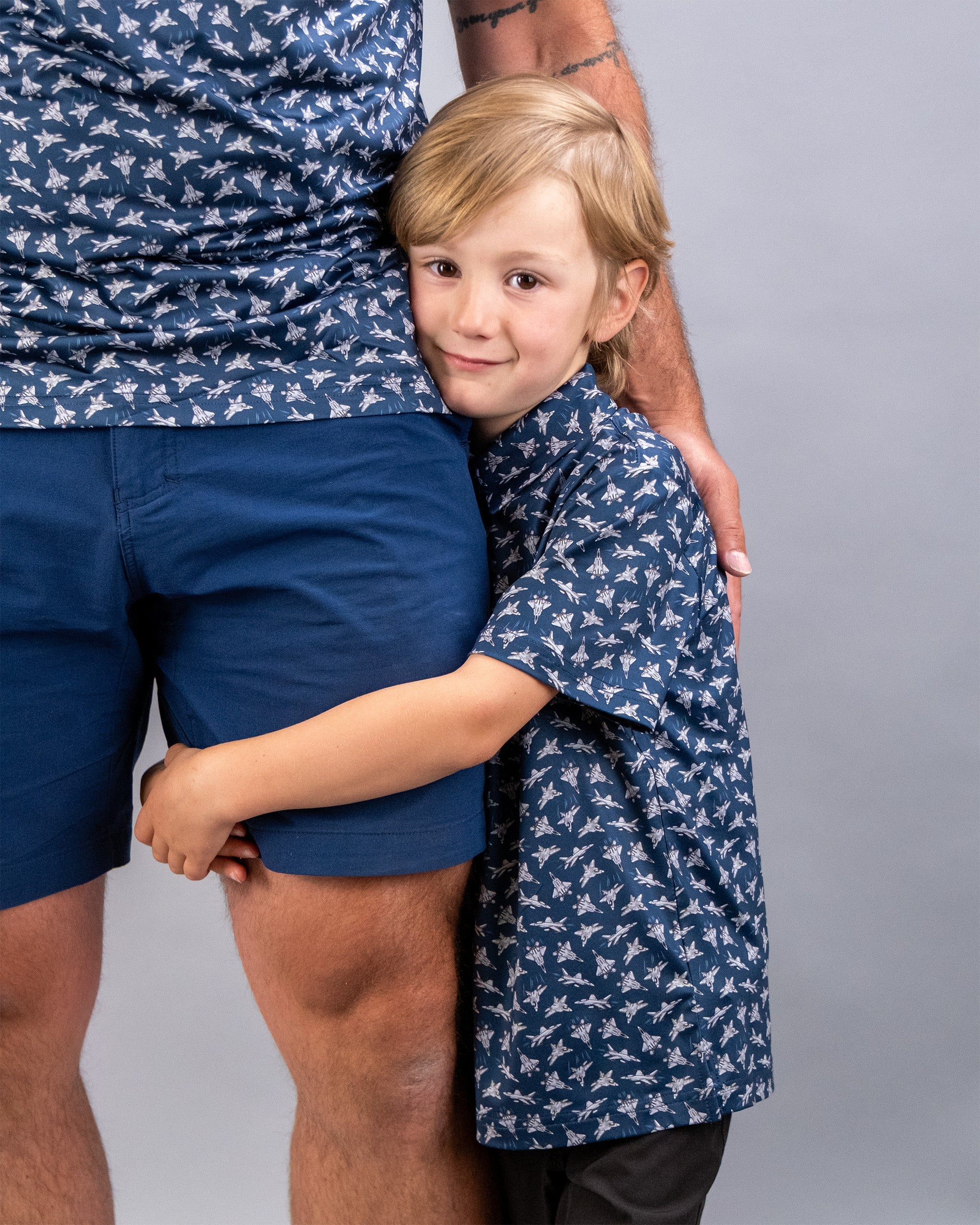 A young boy with blonde hair hugs an adult's leg, both wearing matching blue shirts with a bird pattern, against a plain gray background.