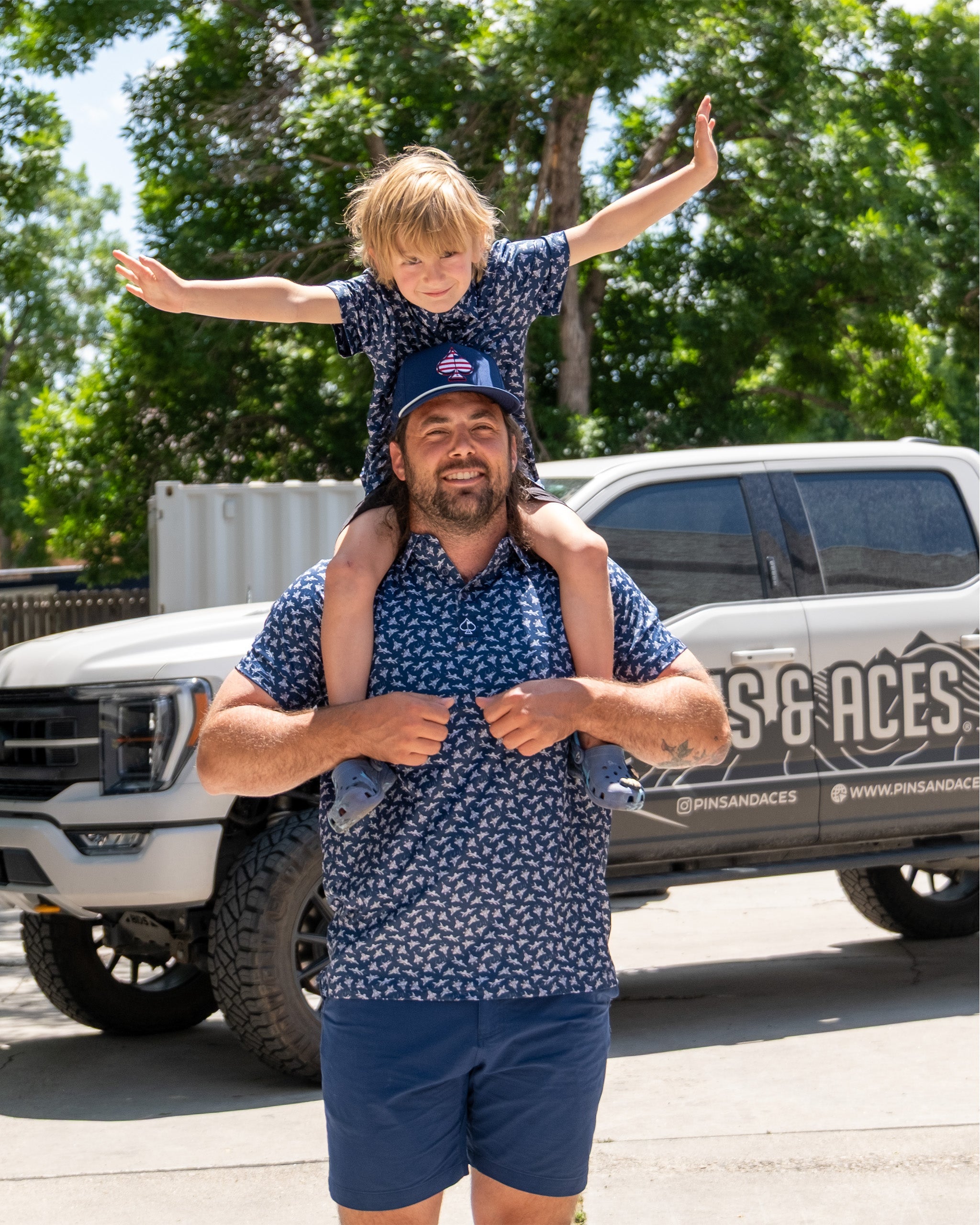 A man with long hair and a beard carries a young boy on his shoulders, both wearing matching blue patterned shirts, outdoors near a vehicle.