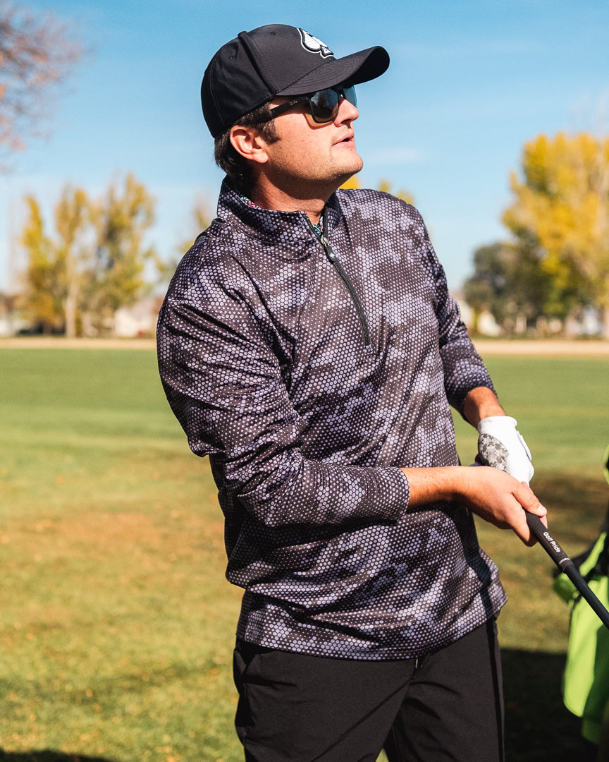 Man wearing a black hex digi pullover, black cap, sunglasses, and holding a golf club outdoors on a sunny day.