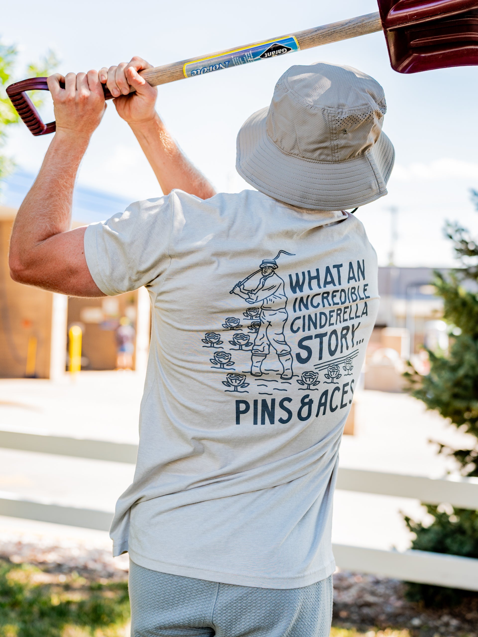 Light beige tee shirt featuring a graphic of a golfer with the text "What an incredible Cinderella story" from Pins and Aces.