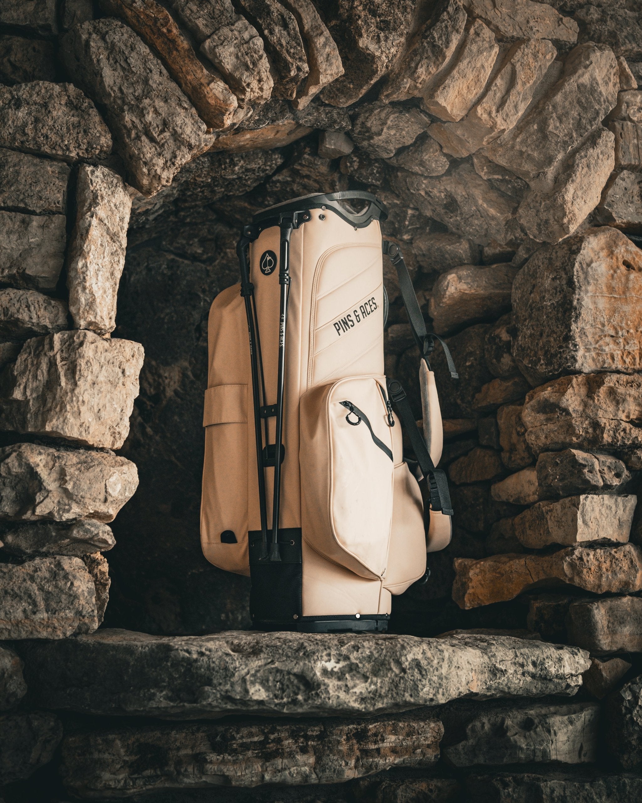Cream-colored golf bag with black accents, labeled "PINS & ACES," surrounded by rustic stone wall, emphasizing bold, uniqu...