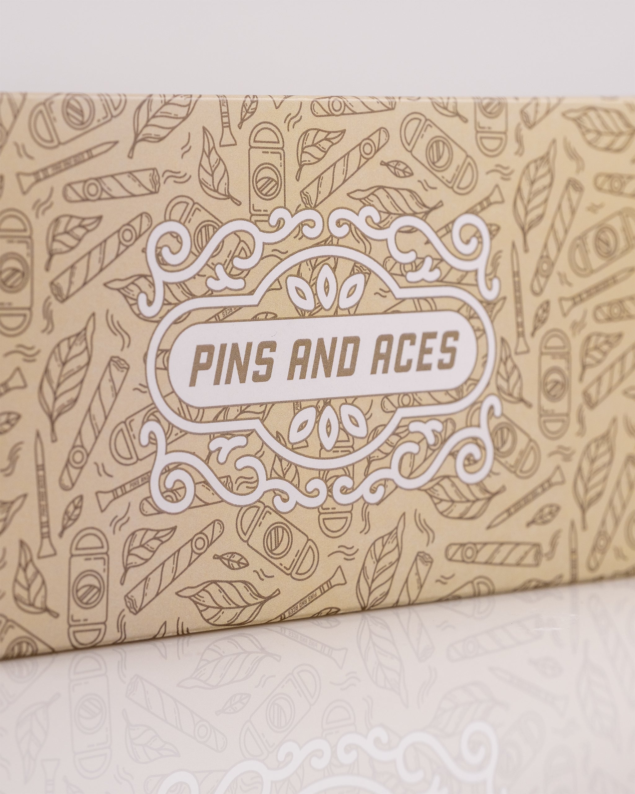 Beige box with "Pins and Aces" logo, decorated with playful golf-themed illustrations like tees, leaves, and golf accessor...