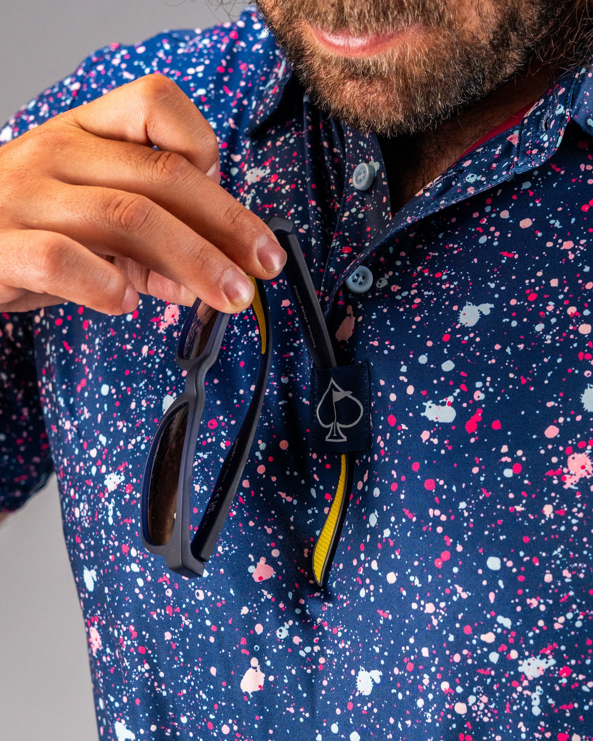 A golfer wearing a blue splatter polo shirt holds sunglasses near the collar, showcasing a stylish and bold golf look.