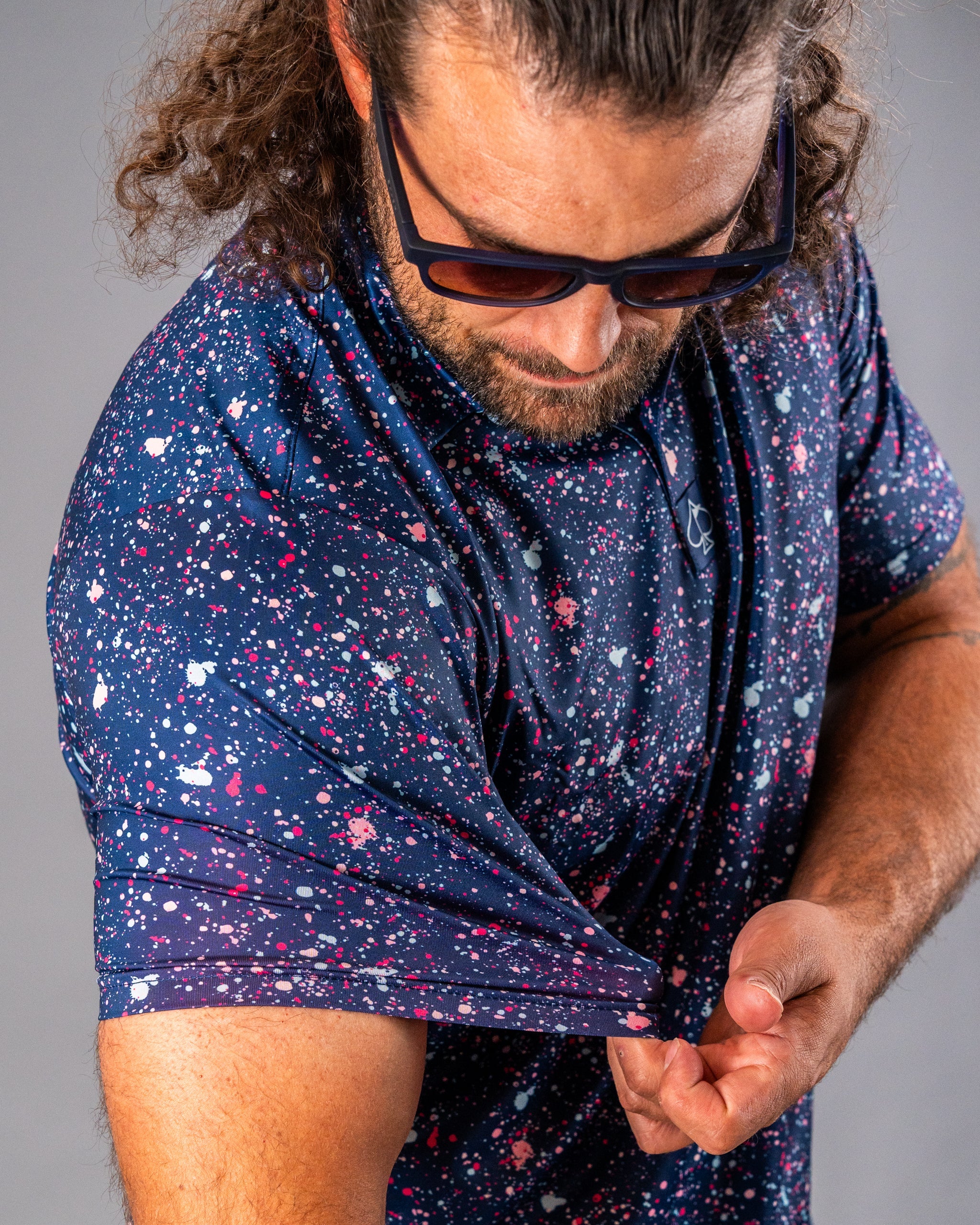A golfer adjusts the sleeve of a blue splatter polo shirt, showcasing its vibrant, colorful design against a gray background.