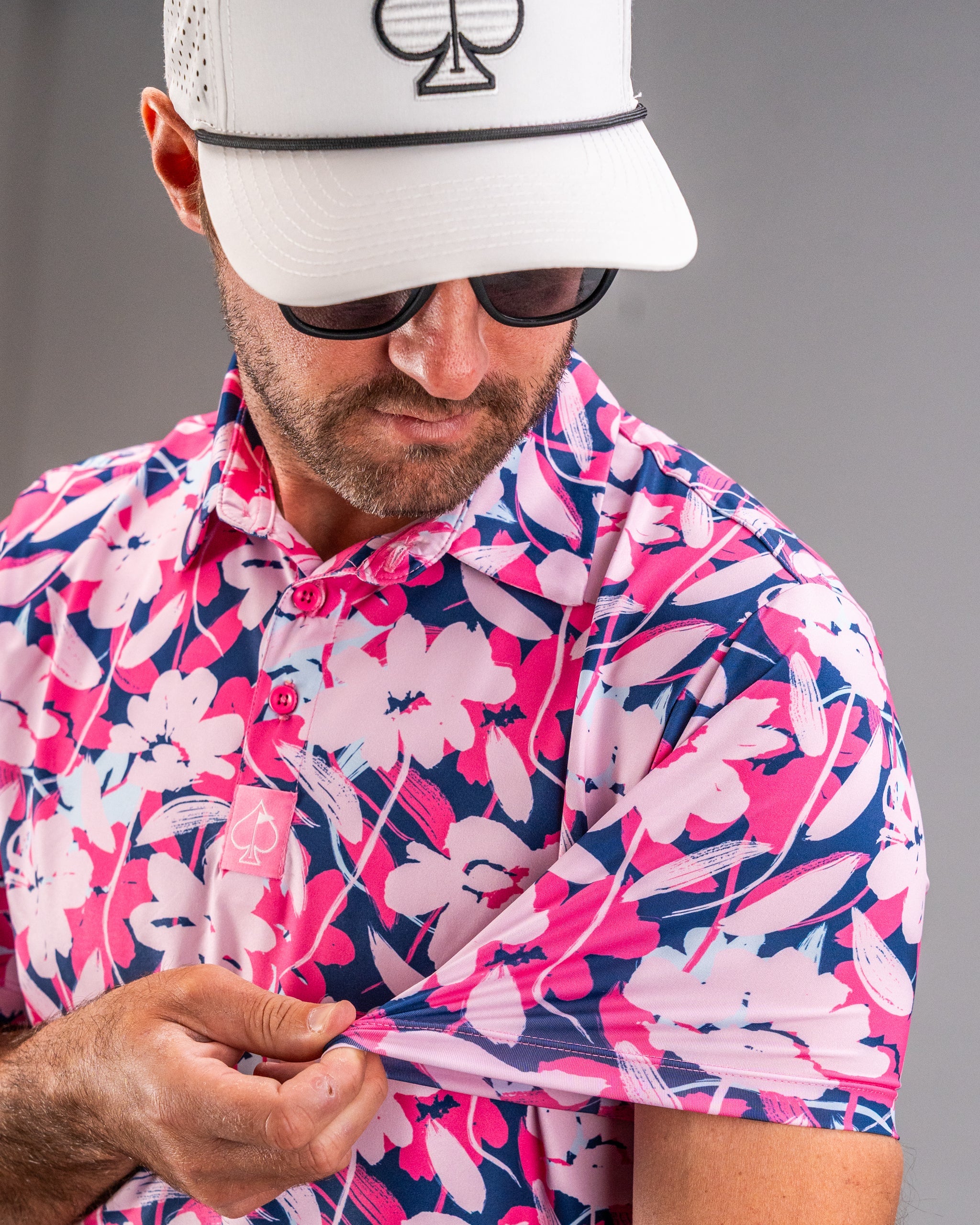Man wearing a pink, navy, and white floral golf shirt, adjusting his sleeve, with a white cap and black sunglasses.