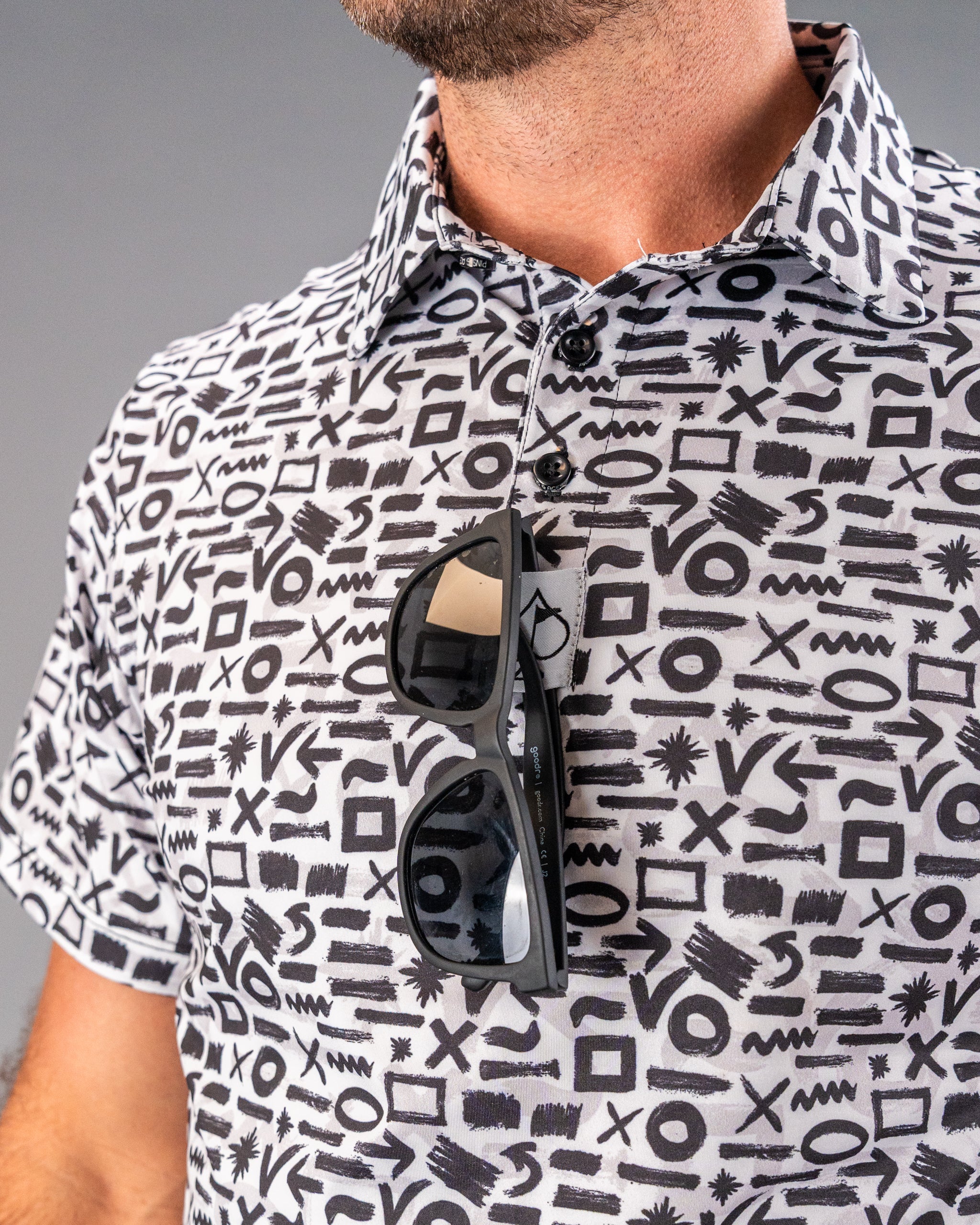 Close-up of a man wearing a bold black-and-white patterned golf shirt with abstract symbols, with sunglasses hanging from ...