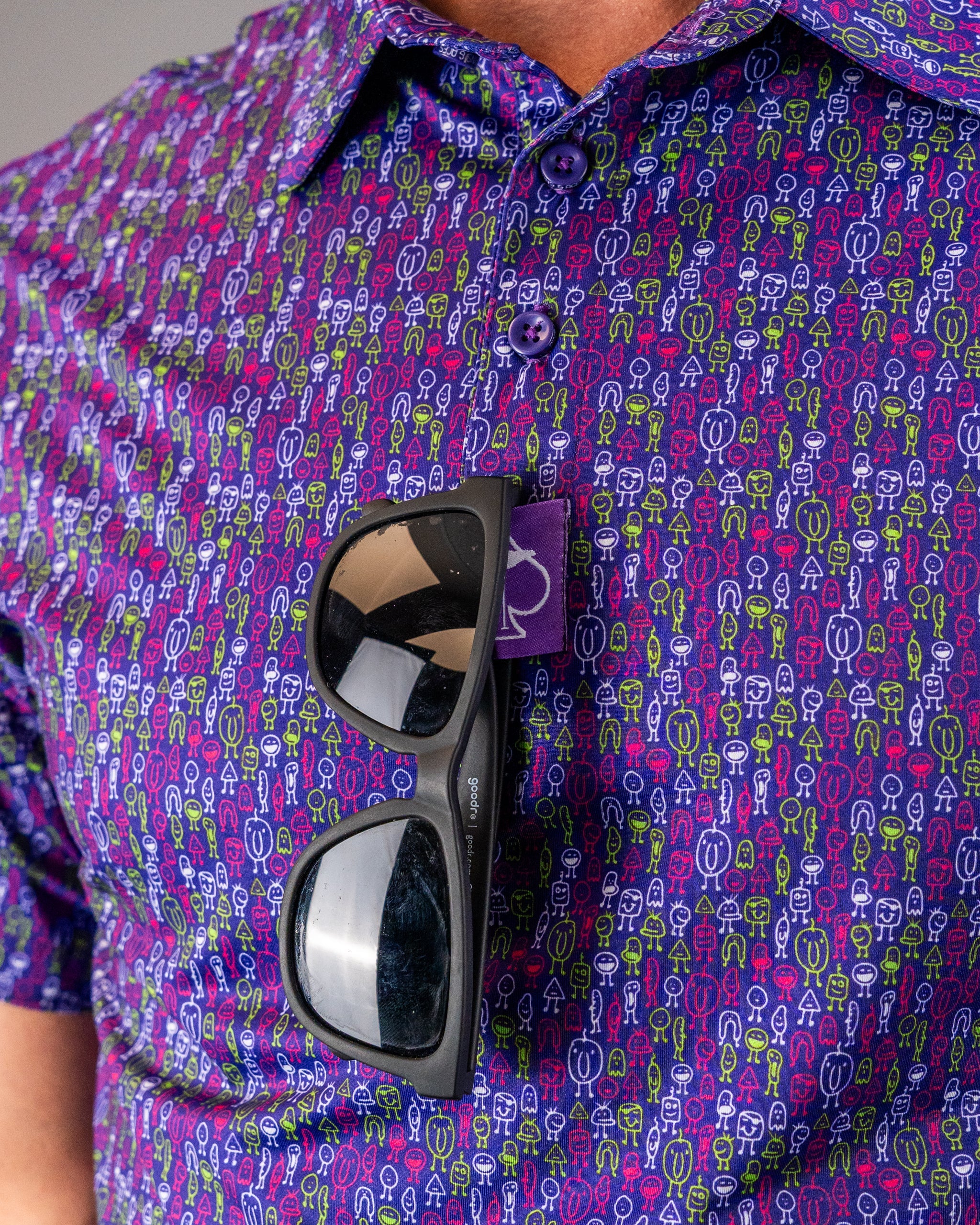 Bright purple shirt with multicolored abstract smiley face pattern, sunglasses tucked in front pocket, and purple buttons.