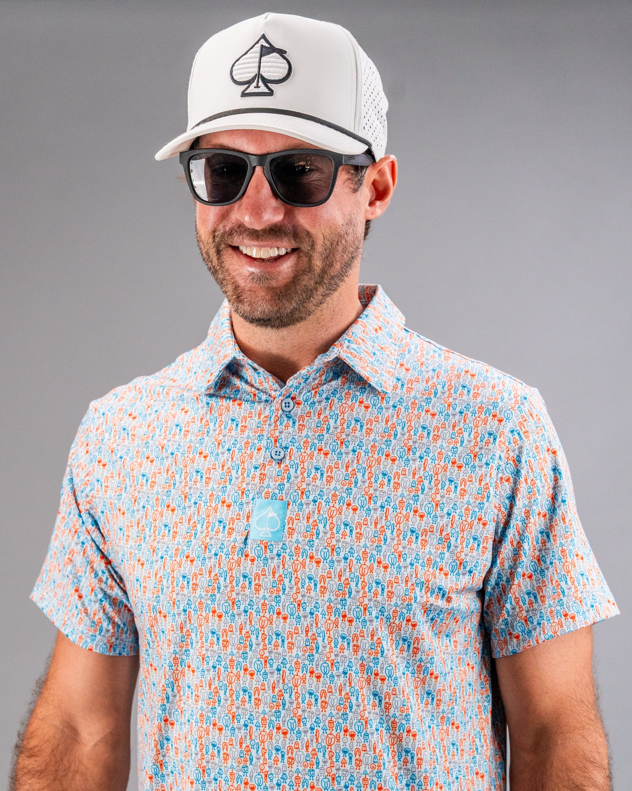 Man wearing a white golf cap with a black spade logo, black sunglasses, and a colorful patterned golf shirt in orange, blu...