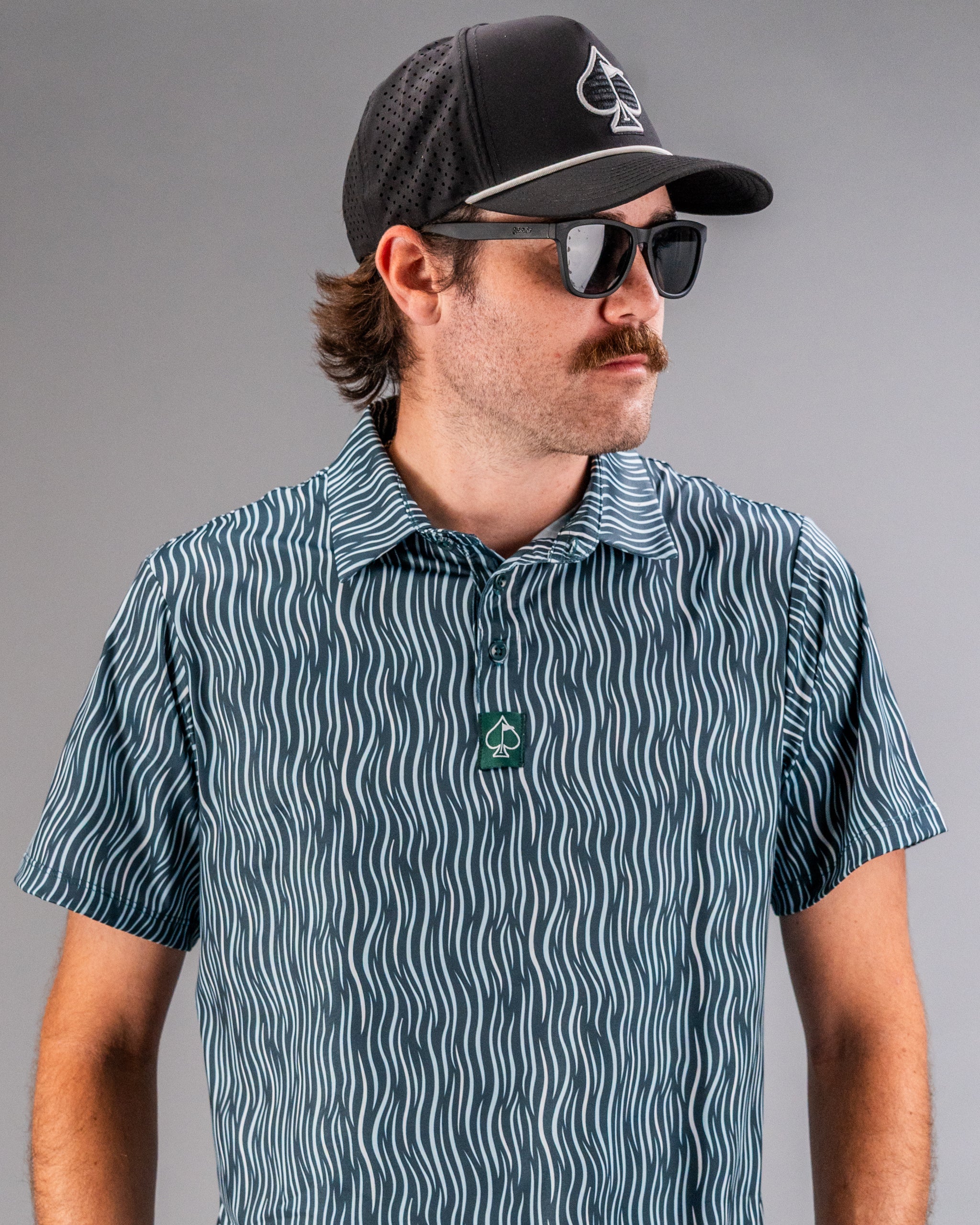 Man wearing a black spade logo cap, dark sunglasses, and a blue and white patterned golf shirt with a green spade logo.