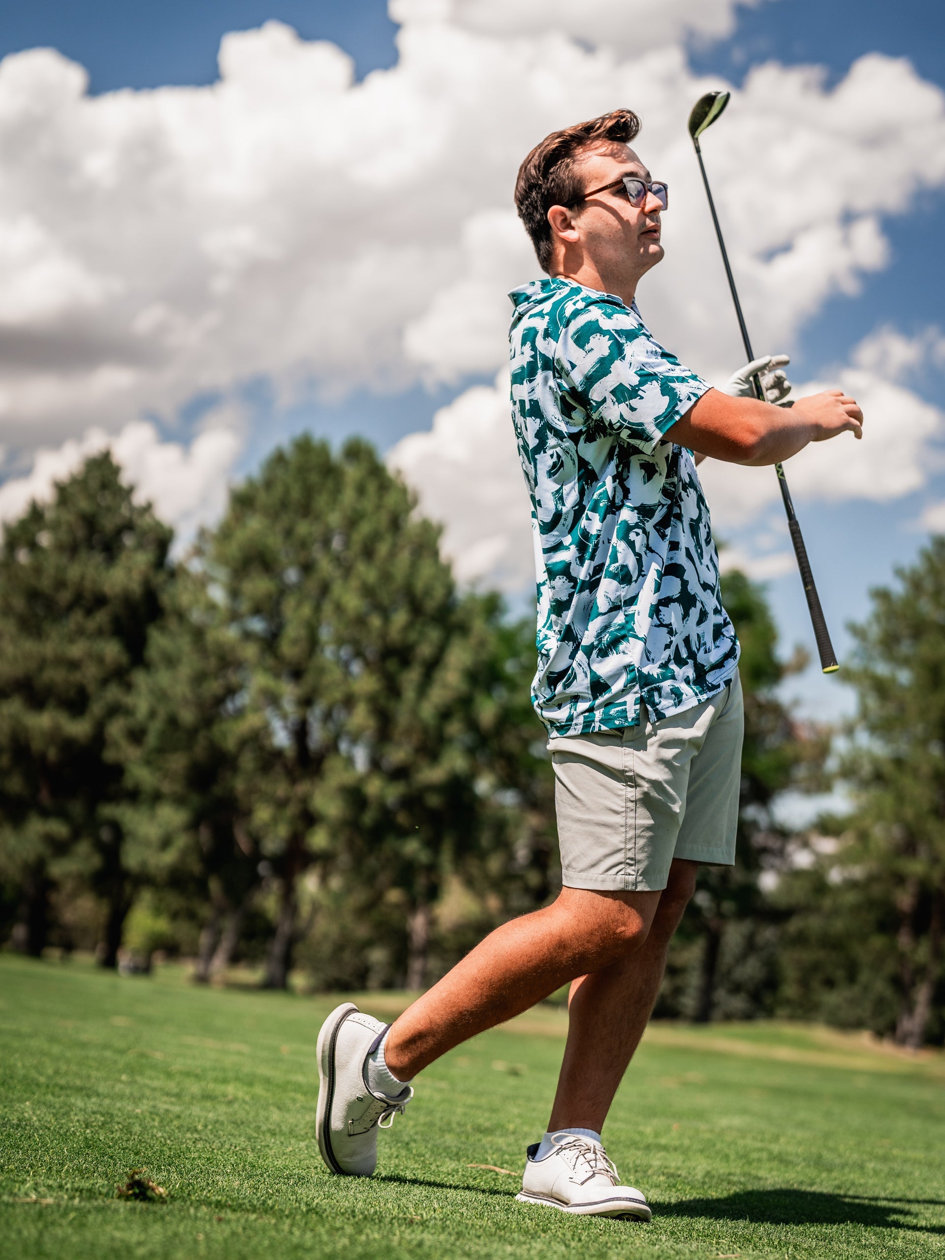 A golfer in a vibrant green paint strokes patterned shirt swings a club on a sunny course, showcasing bold style.