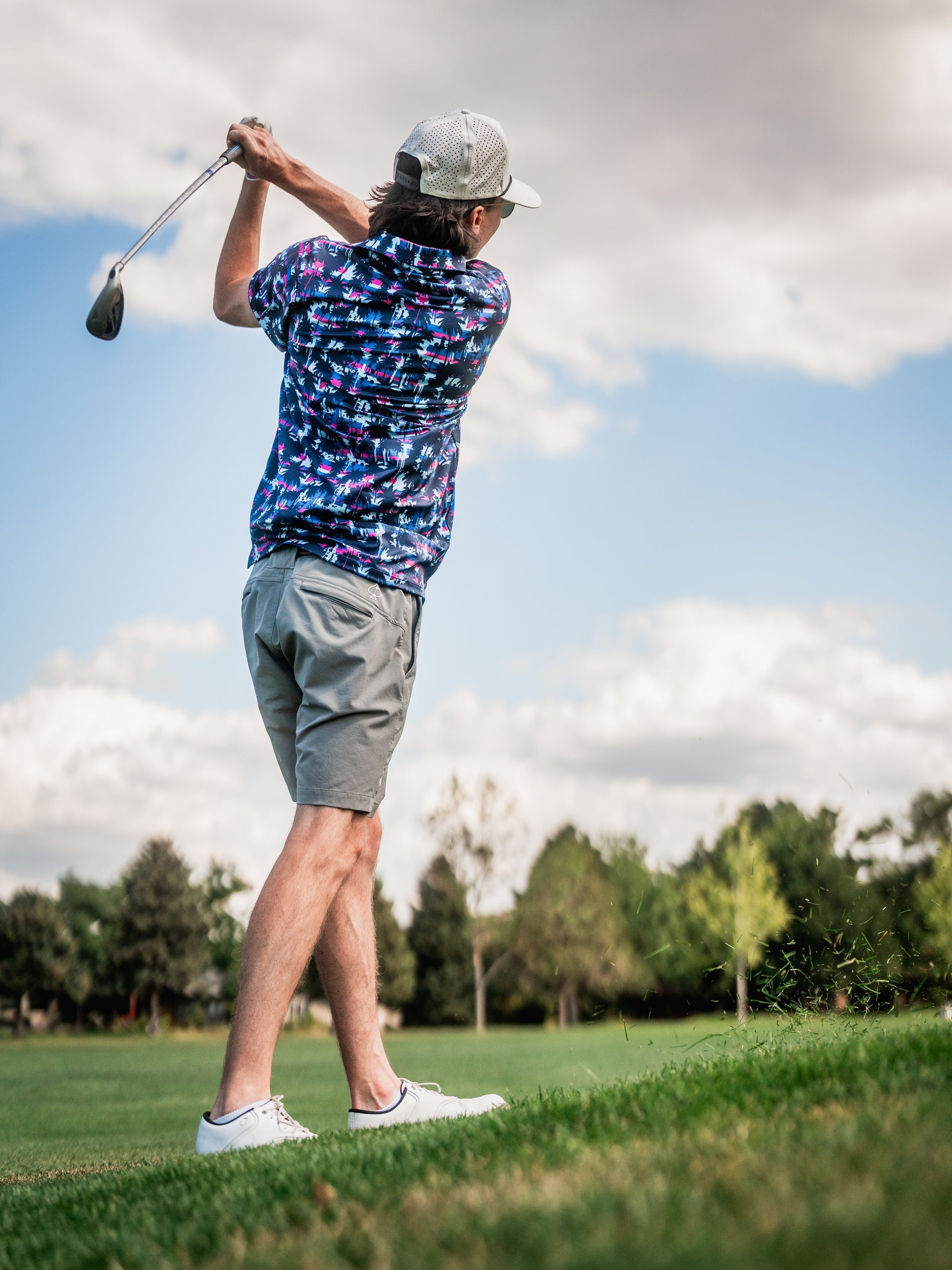 Male golfer in a colorful, patterned shirt, beige shorts, white sneakers, and a white cap, swinging a golf club on a sunny...