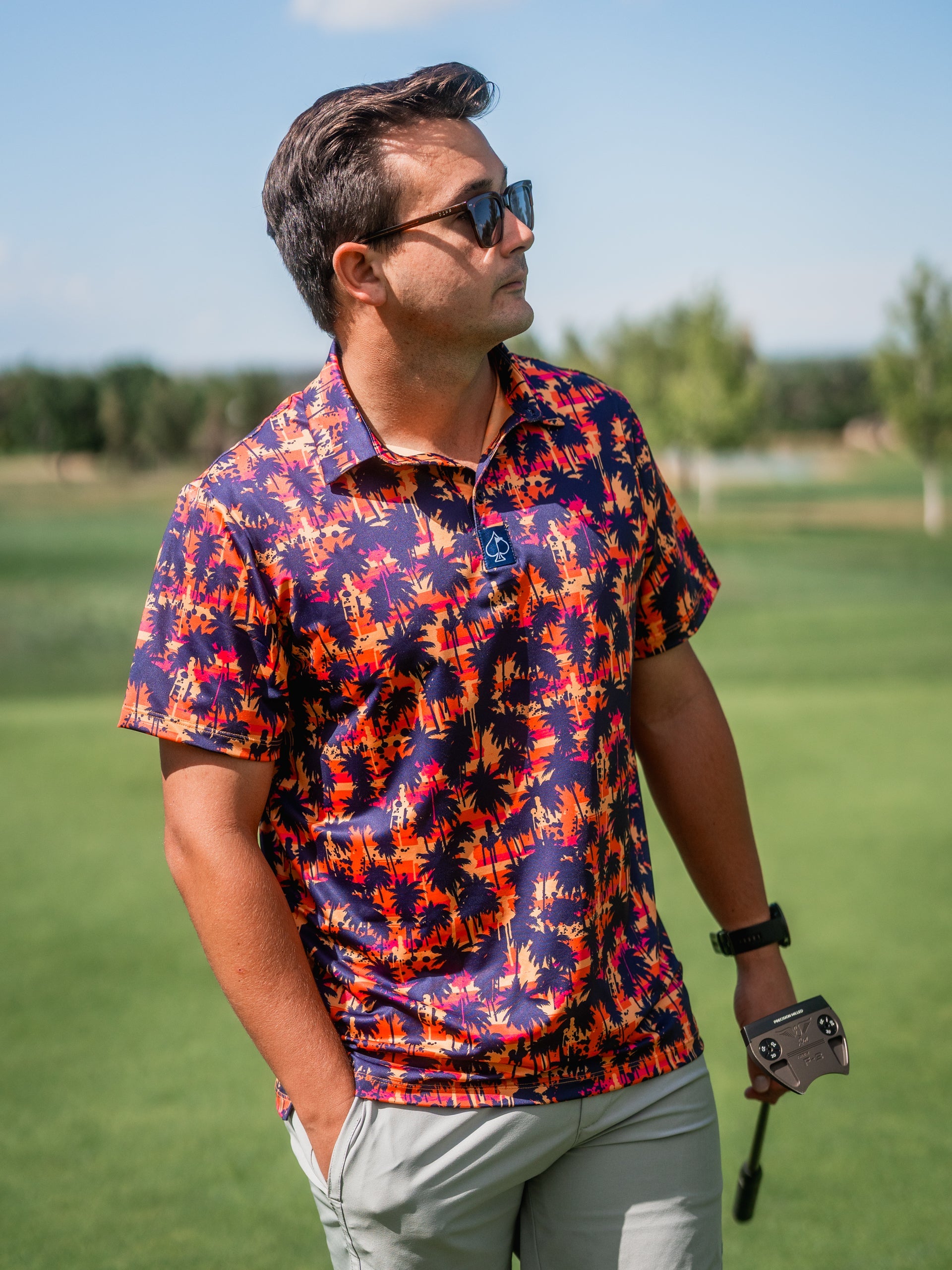 Man wearing a colorful palm print golf shirt, sunglasses, and holding a golf club on the course, showcasing bold golf appa...