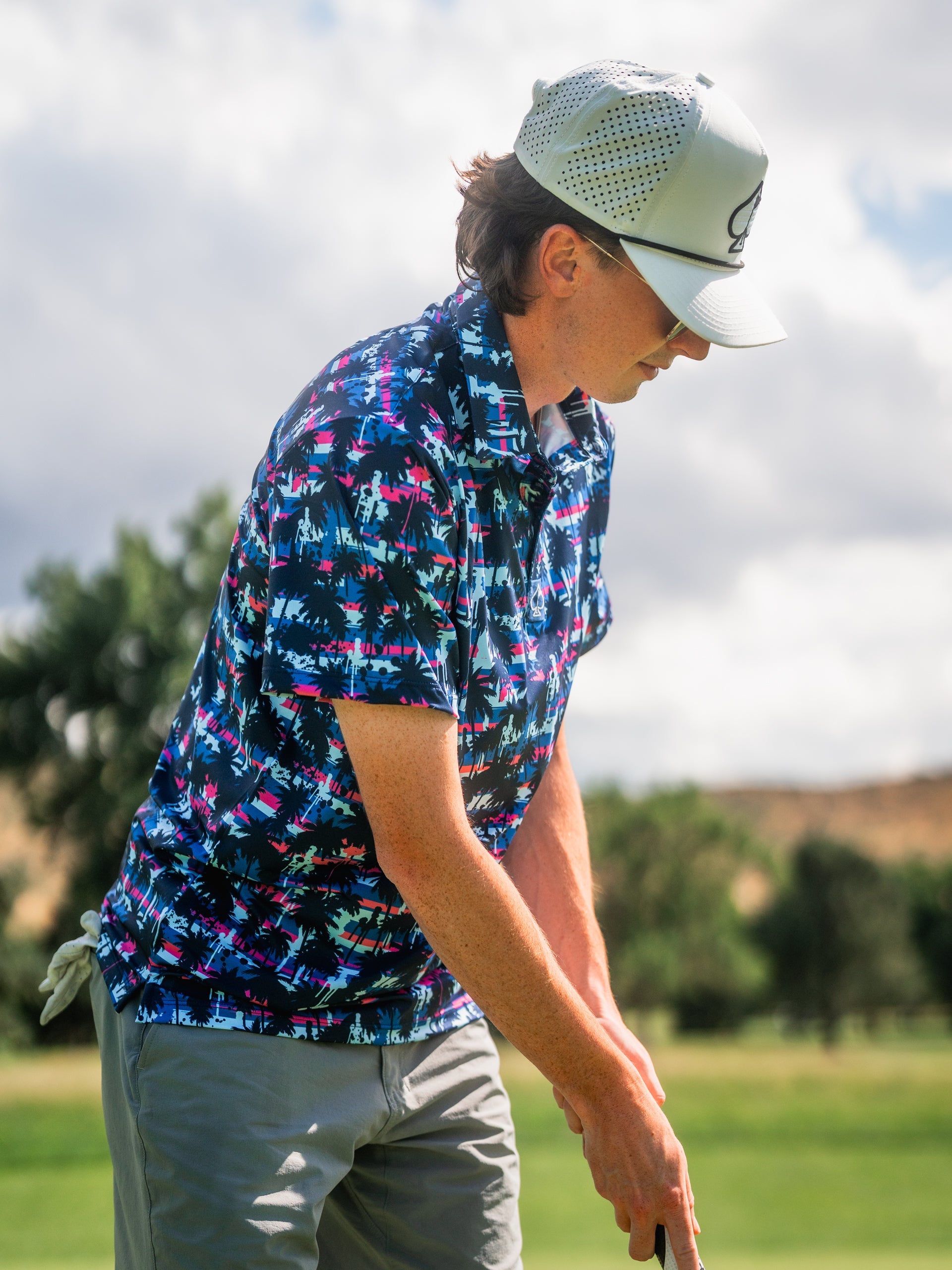 Male golfer in a vibrant tropical-print shirt, white cap, and beige shorts on the course, ready to swing.