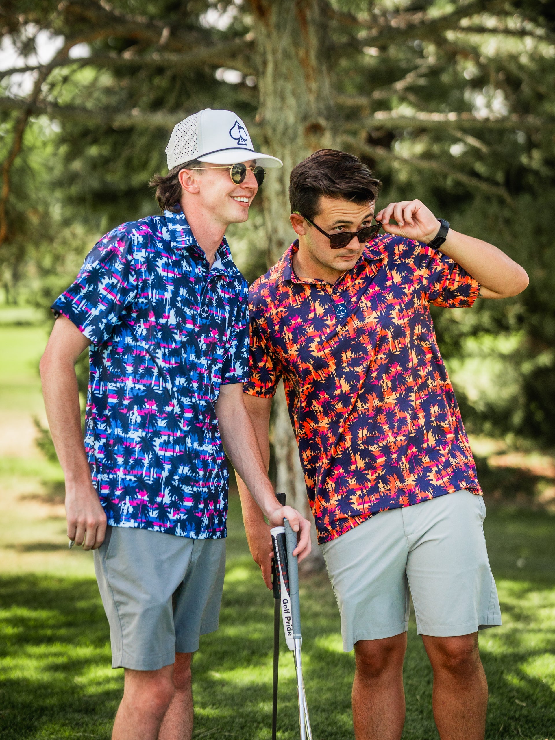 Two men in colorful, bold tropical print golf shirts, light shorts, sunglasses, and hats on a golf course, enjoying a sunn...