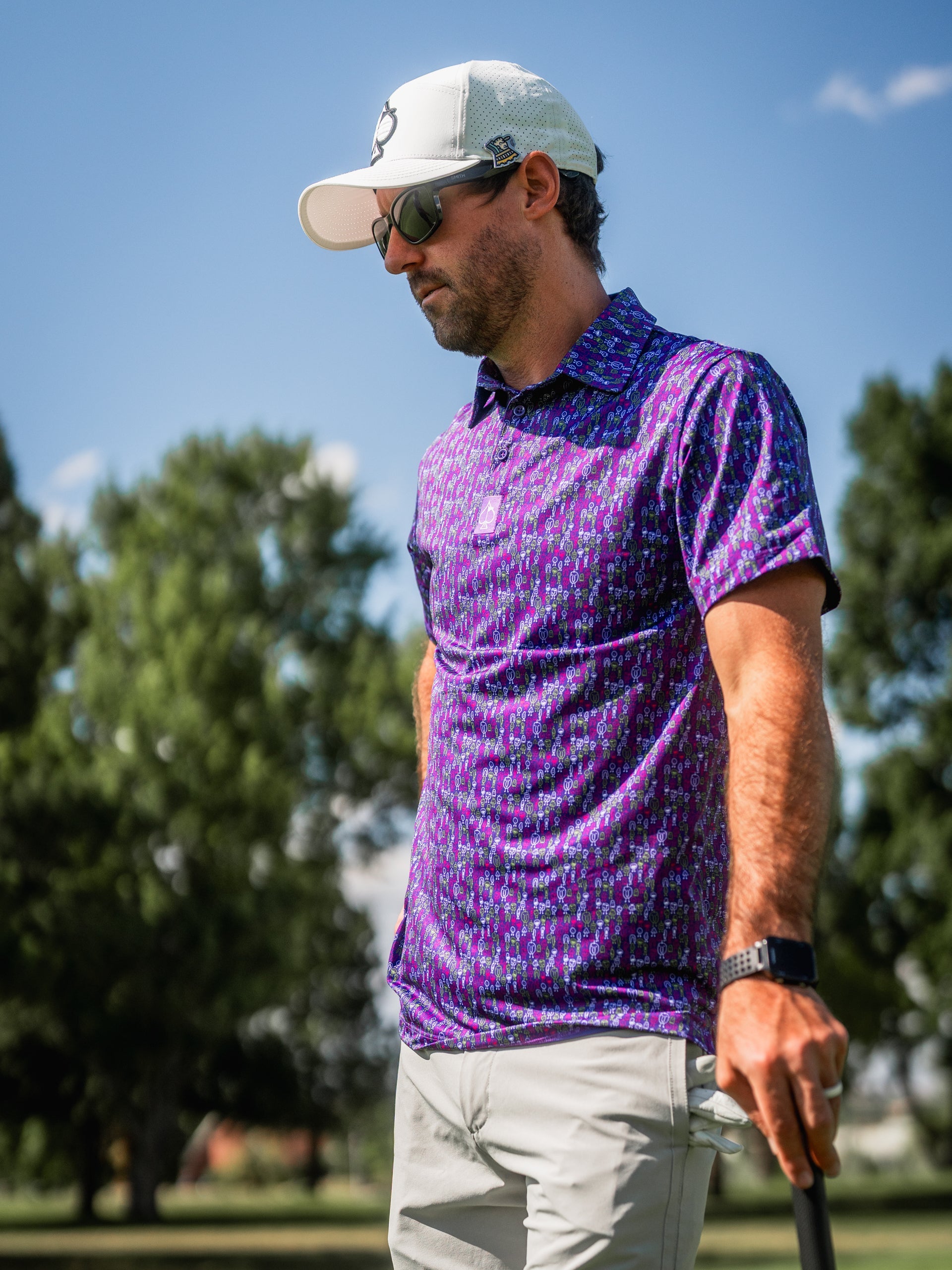 Man in colorful purple and blue golf shirt, white cap, and sunglasses on golf course, showcasing bold golf apparel.