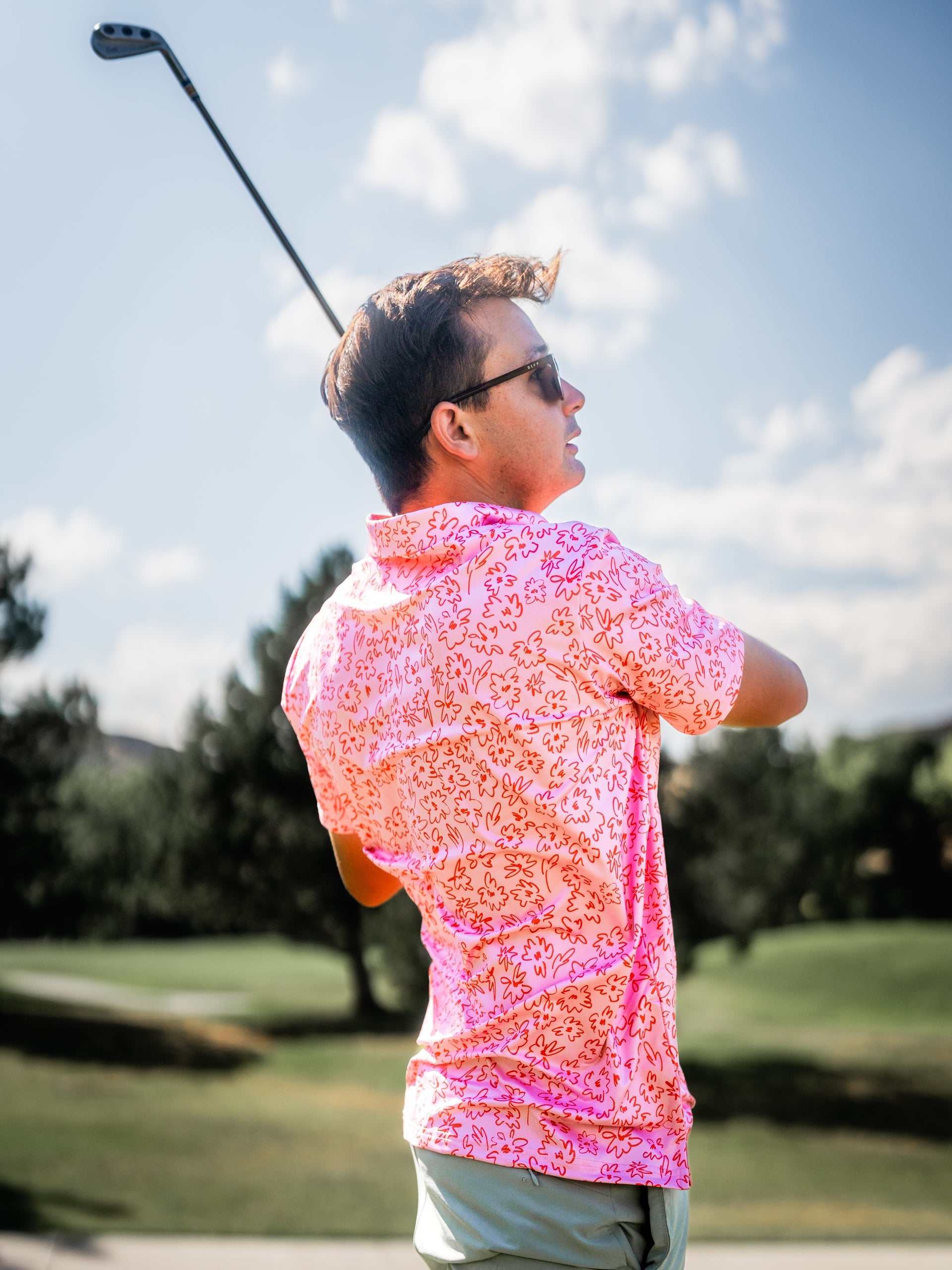 A golfer swings a club while wearing a vibrant pink shirt featuring a floral doodle pattern, set against a sunny golf course.