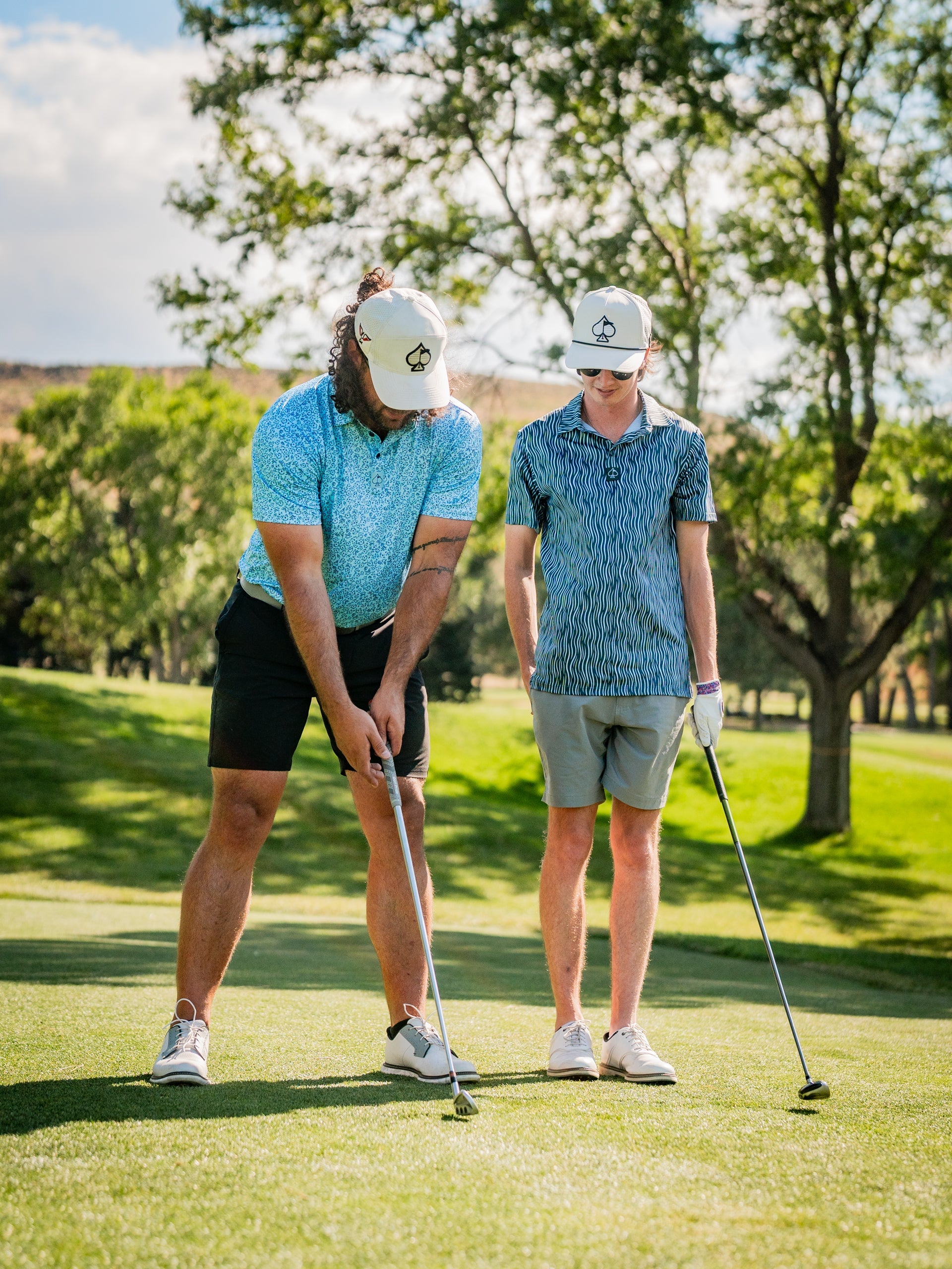 Two golfers on the course, one in a blue flower doodle shirt and the other in a patterned shirt, preparing to putt.