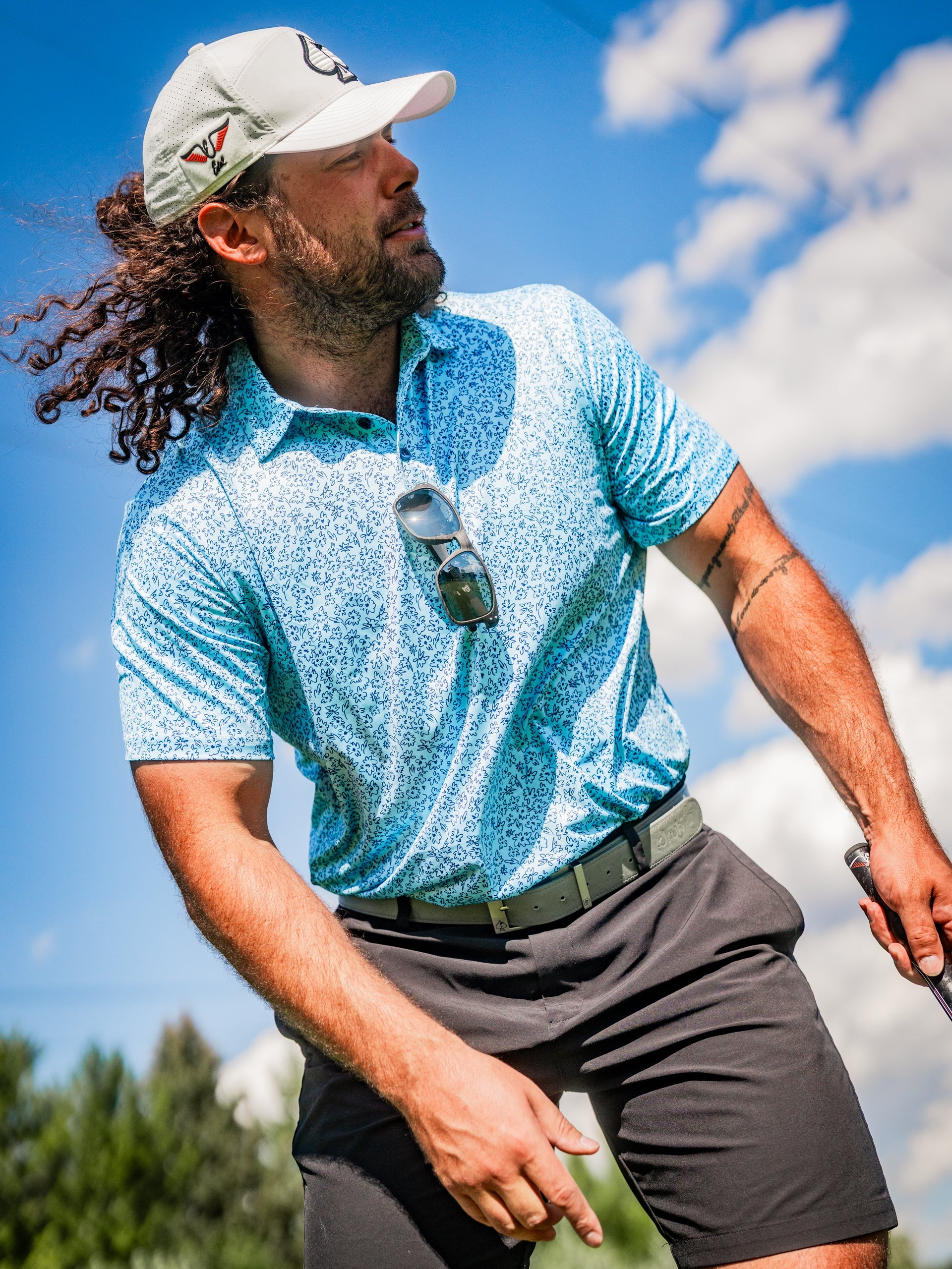 A golfer wearing a blue flower doodle polo shirt and gray shorts, preparing to swing on a sunny day.