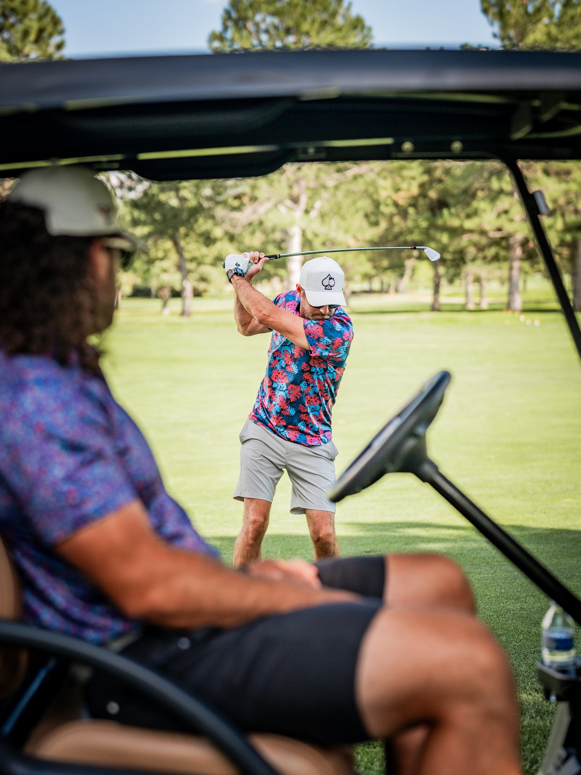 Two golfers on a lush green course, one swinging a club, wearing bold, colorful golf apparel; the other relaxing in a golf...