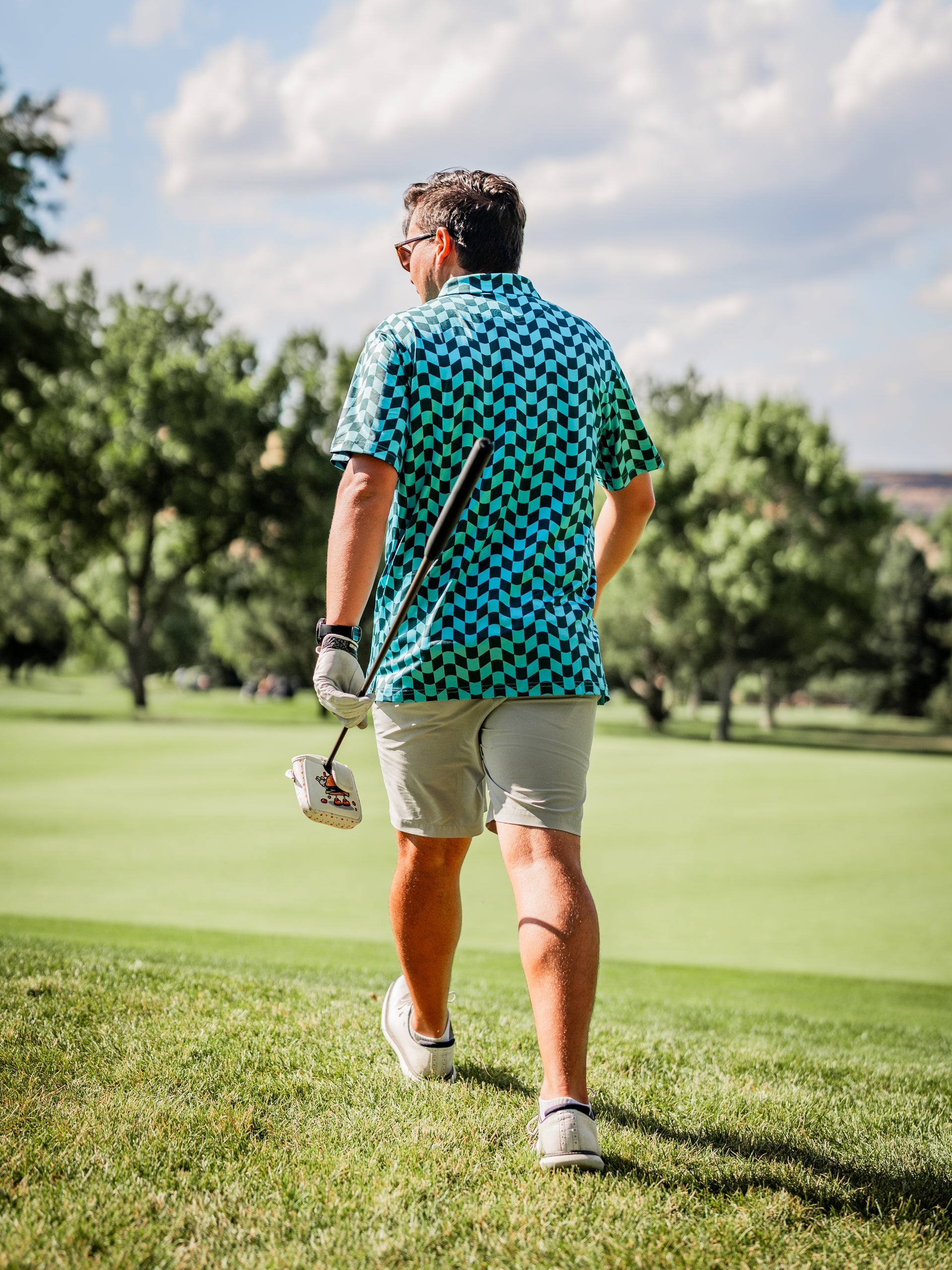 Man walking on a golf course wearing a wavy checkered blue and green polo shirt and shorts, holding a golf club.