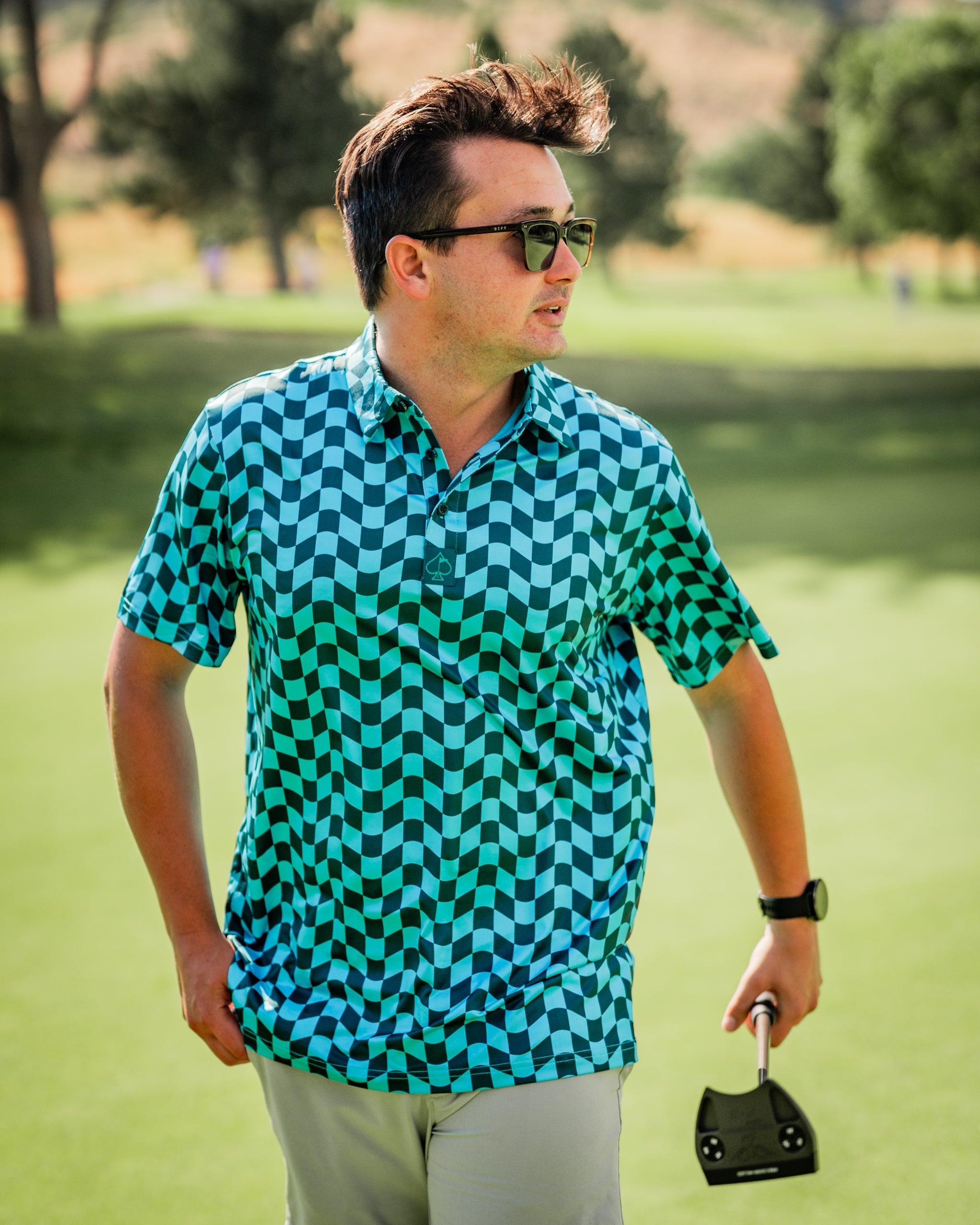 A golfer wearing a vibrant wavy checkered polo shirt in shades of blue and green, walking on the course with a putter.