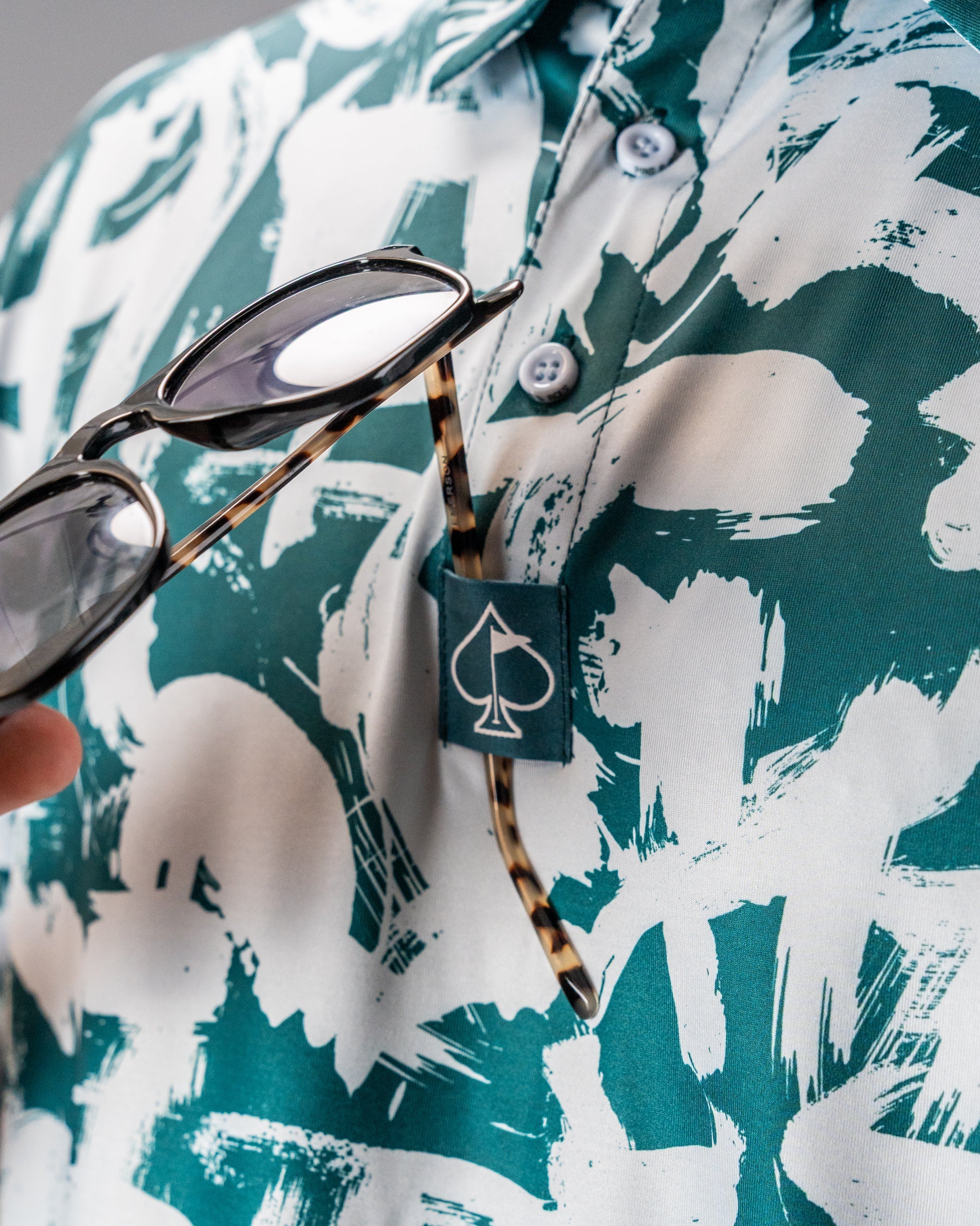 Close-up of a green and white patterned golf shirt with a logo tag, featuring sunglasses resting on the collar.