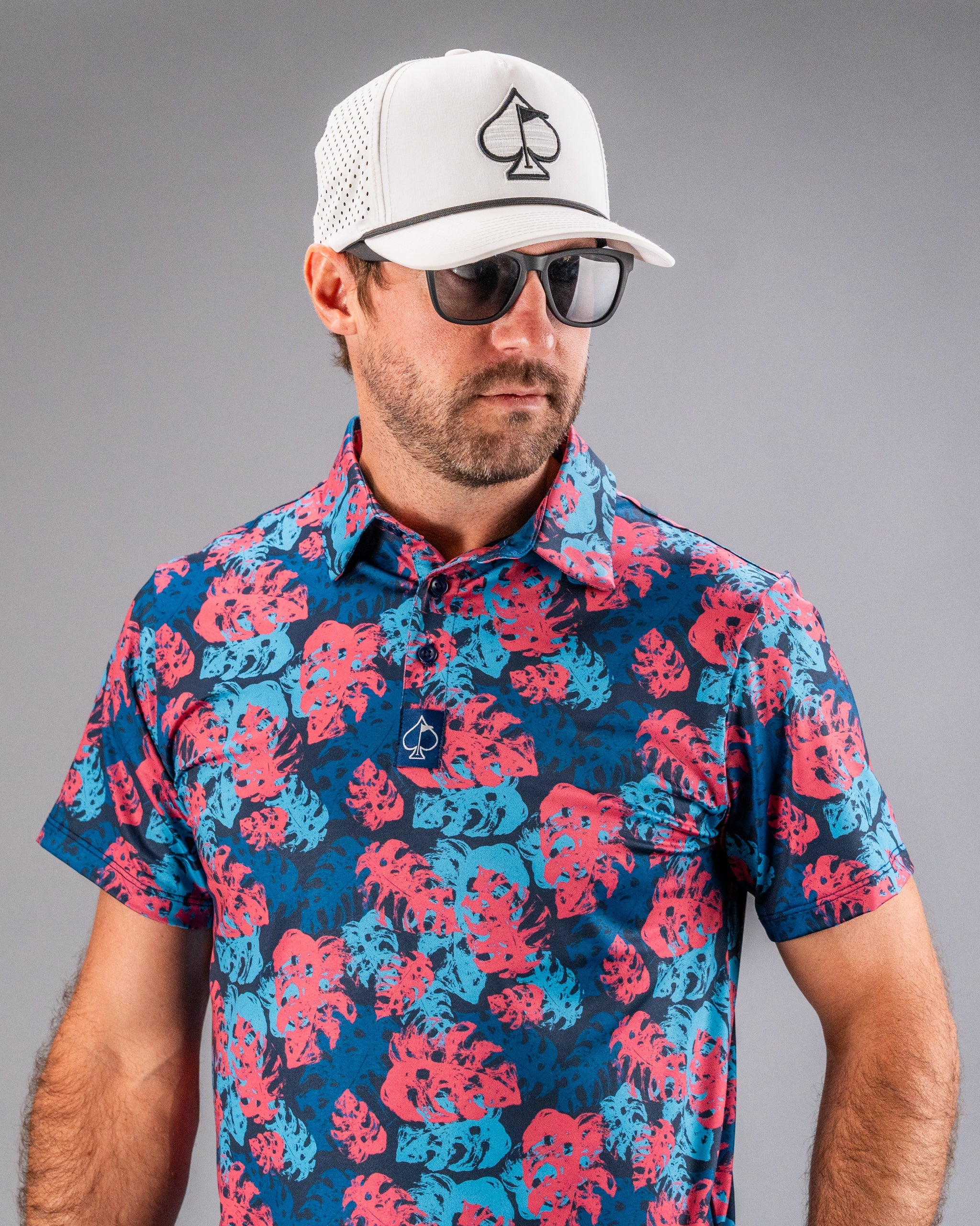 Men's vibrant pink and blue floral golf shirt with a spade logo, paired with a white mesh cap featuring a black spade embl...