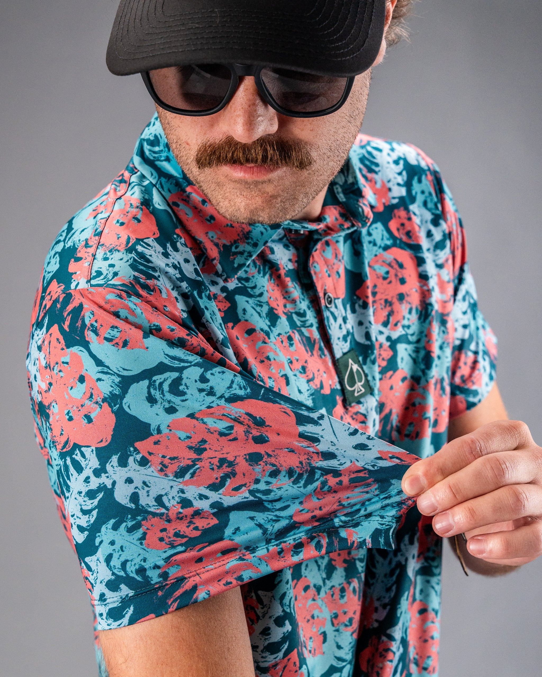 Man in a vibrant blue and coral floral golf shirt, wearing a black cap, sunglasses, and showcasing bold golf apparel.