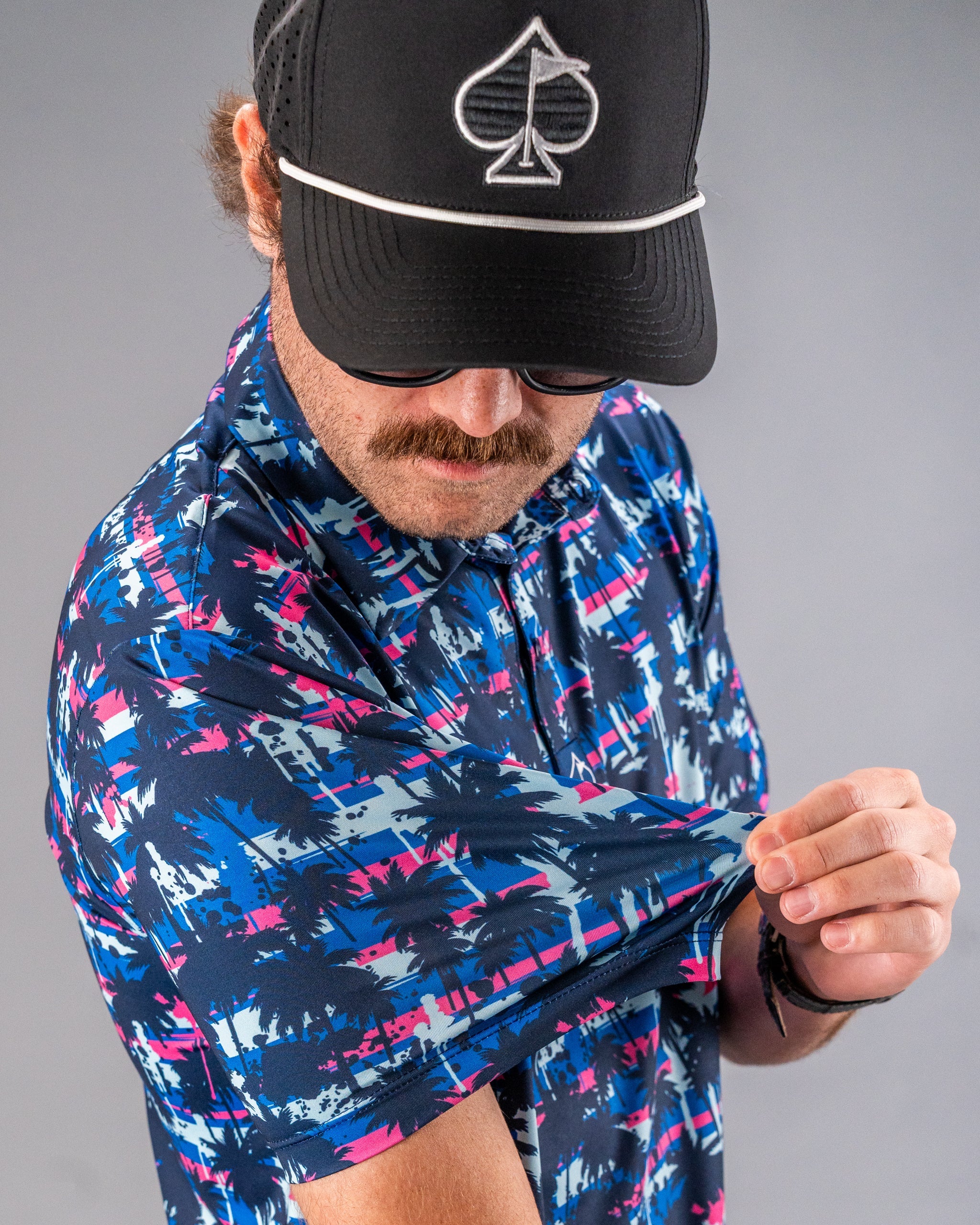 Man in colorful palm tree patterned golf shirt, adjusting sleeve, wearing black cap with spade logo, for bold golf style.