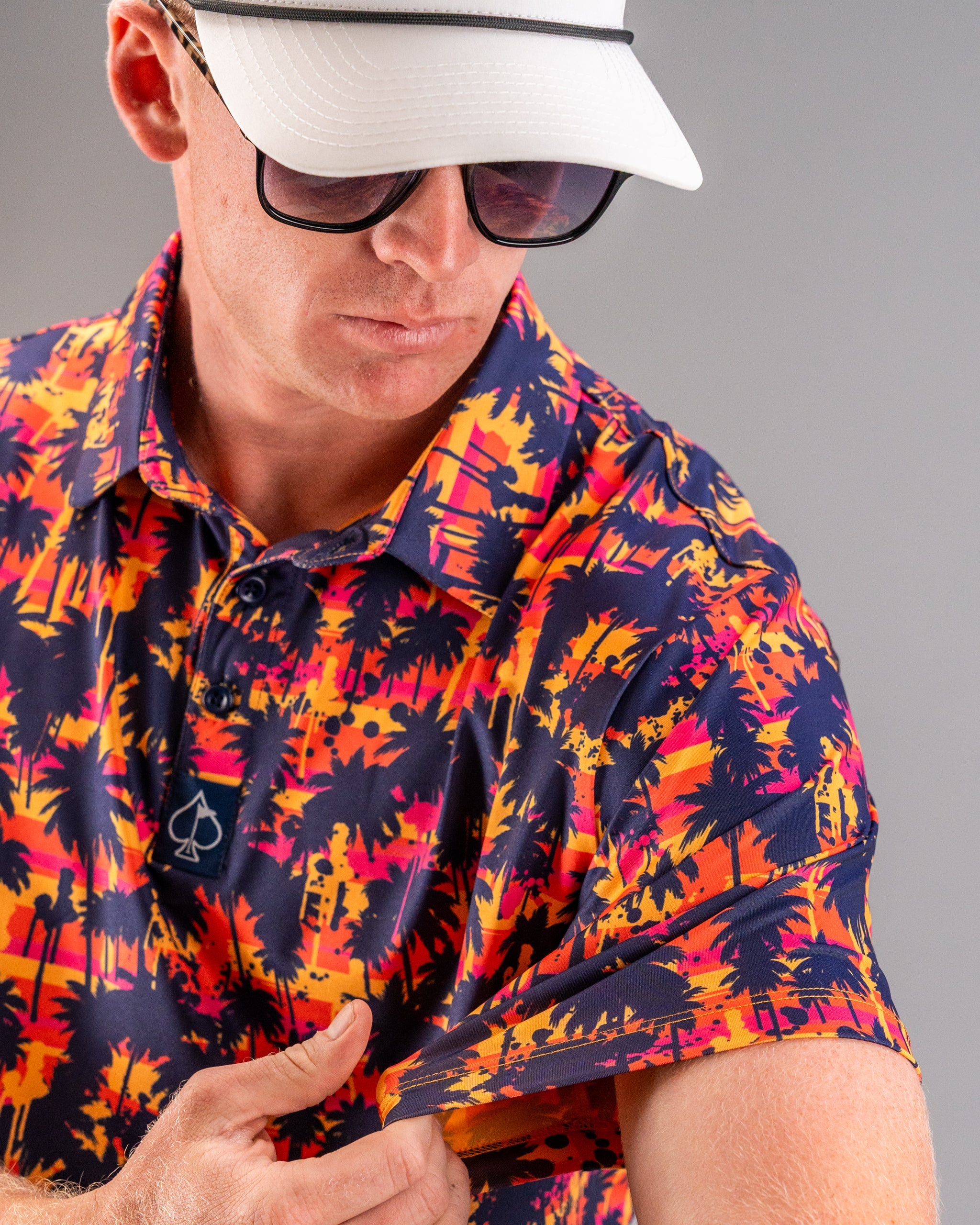 Close-up of a man wearing a vibrant, tropical-print golf shirt in navy, pink, and orange, paired with a white golf cap and...