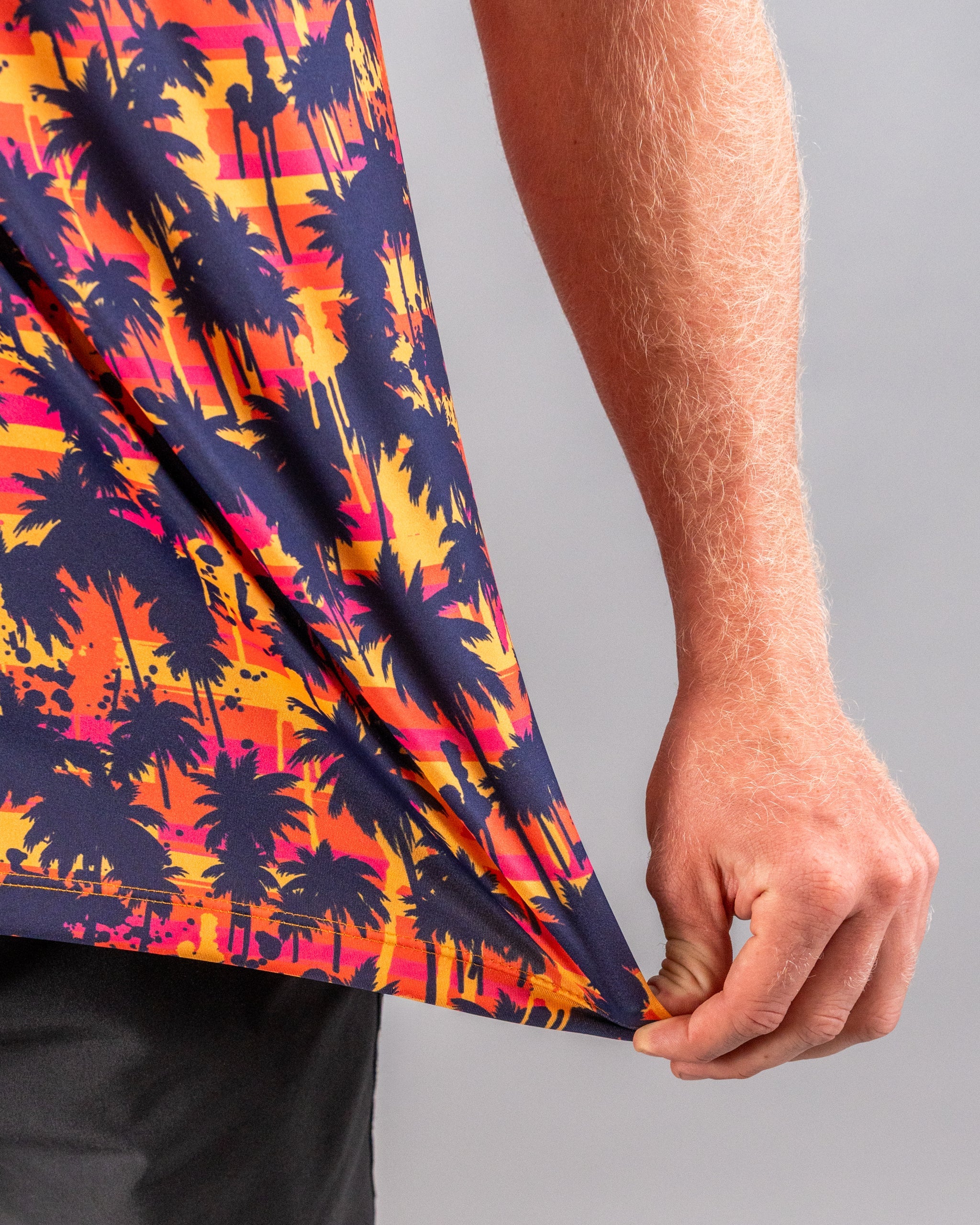Close-up of a colorful golf shirt featuring a vibrant sunset and palm tree print, with a person's hand pulling at the hem.